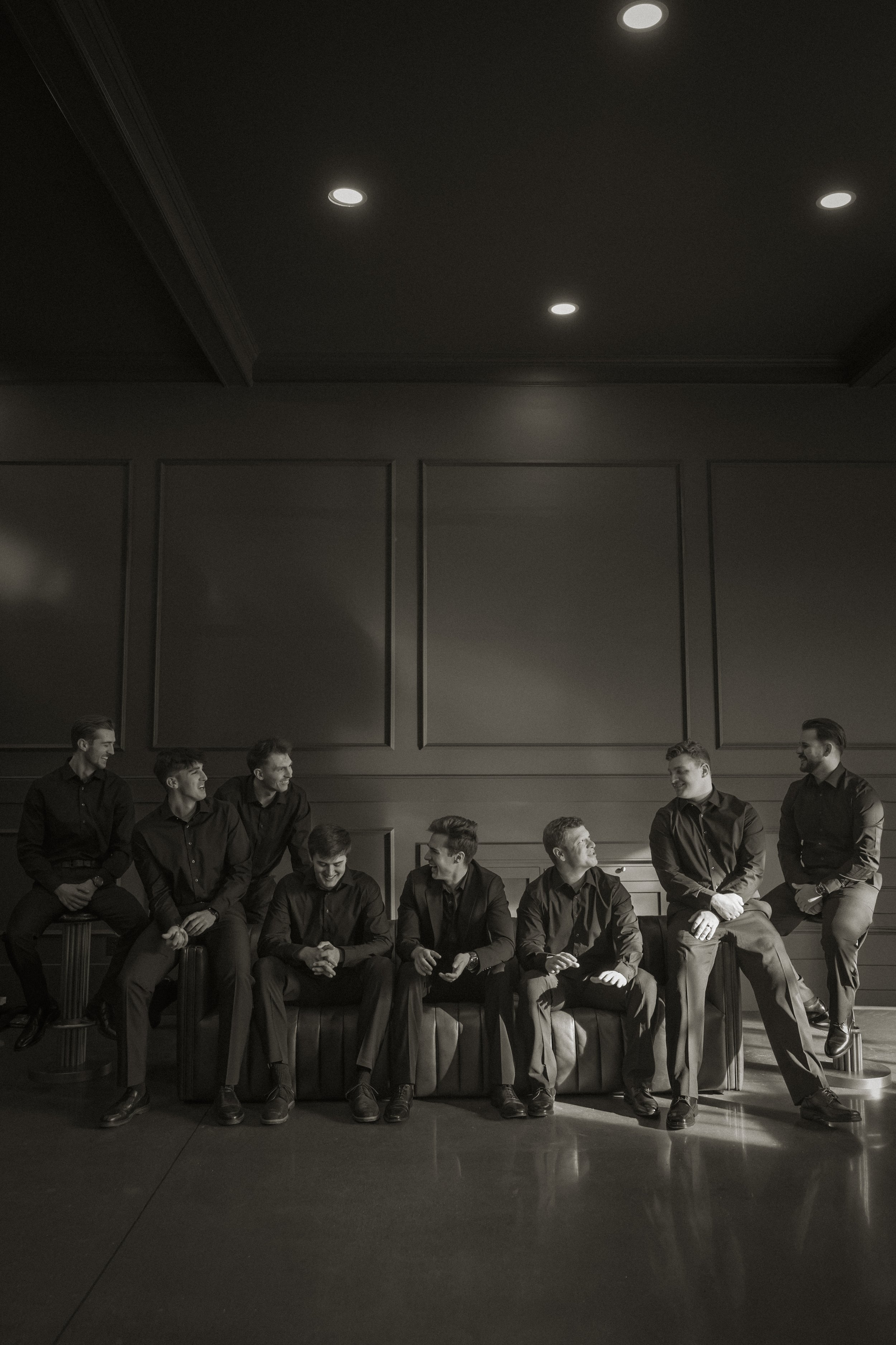 A black and white photo of nine men dressed in formal black shirts and pants, sitting and standing on a bench in a room with a plain wall and ceiling lights.