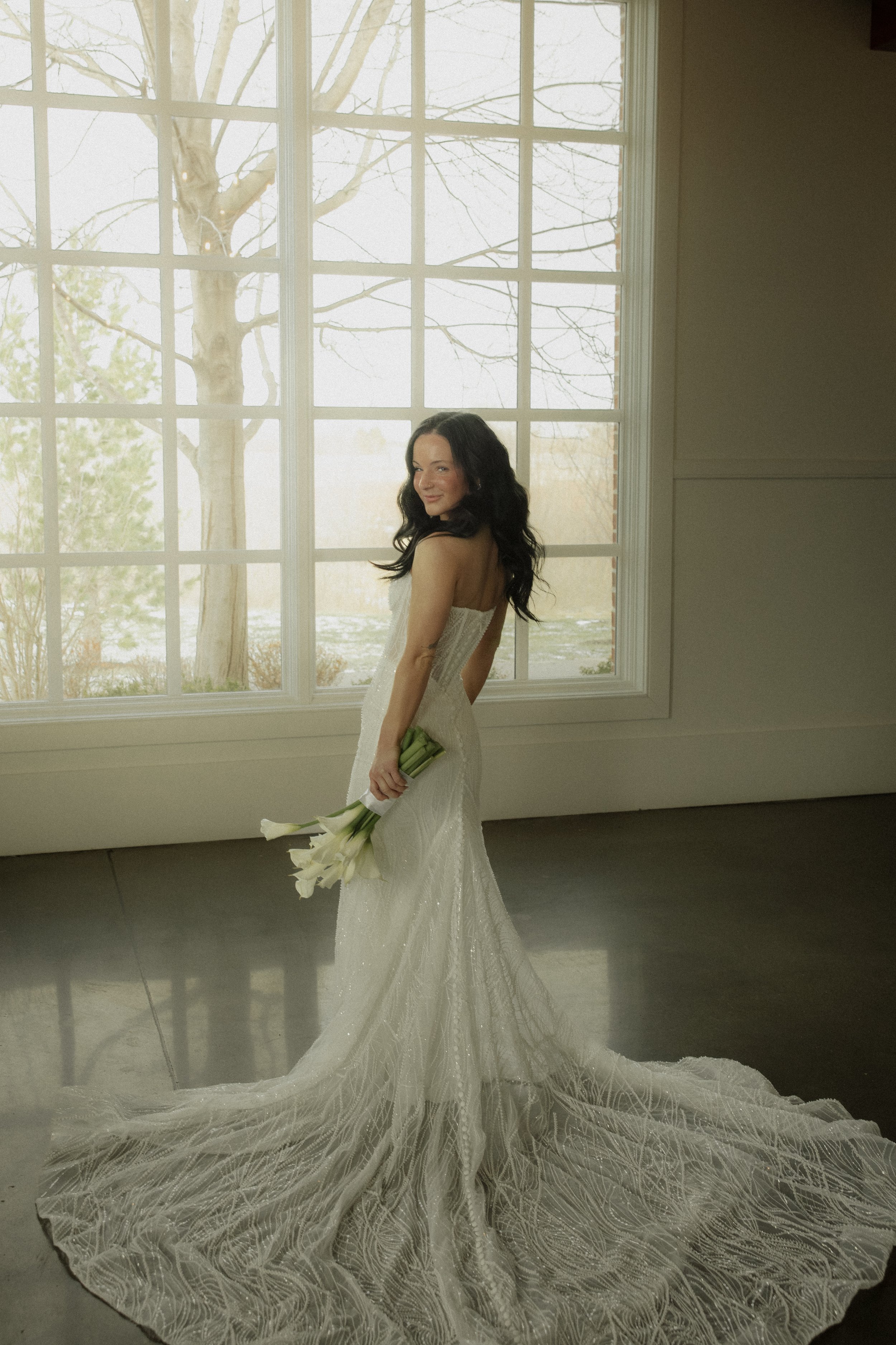 A woman in a white wedding dress holding a bouquet of white calla lilies, standing inside near large window with trees outside.