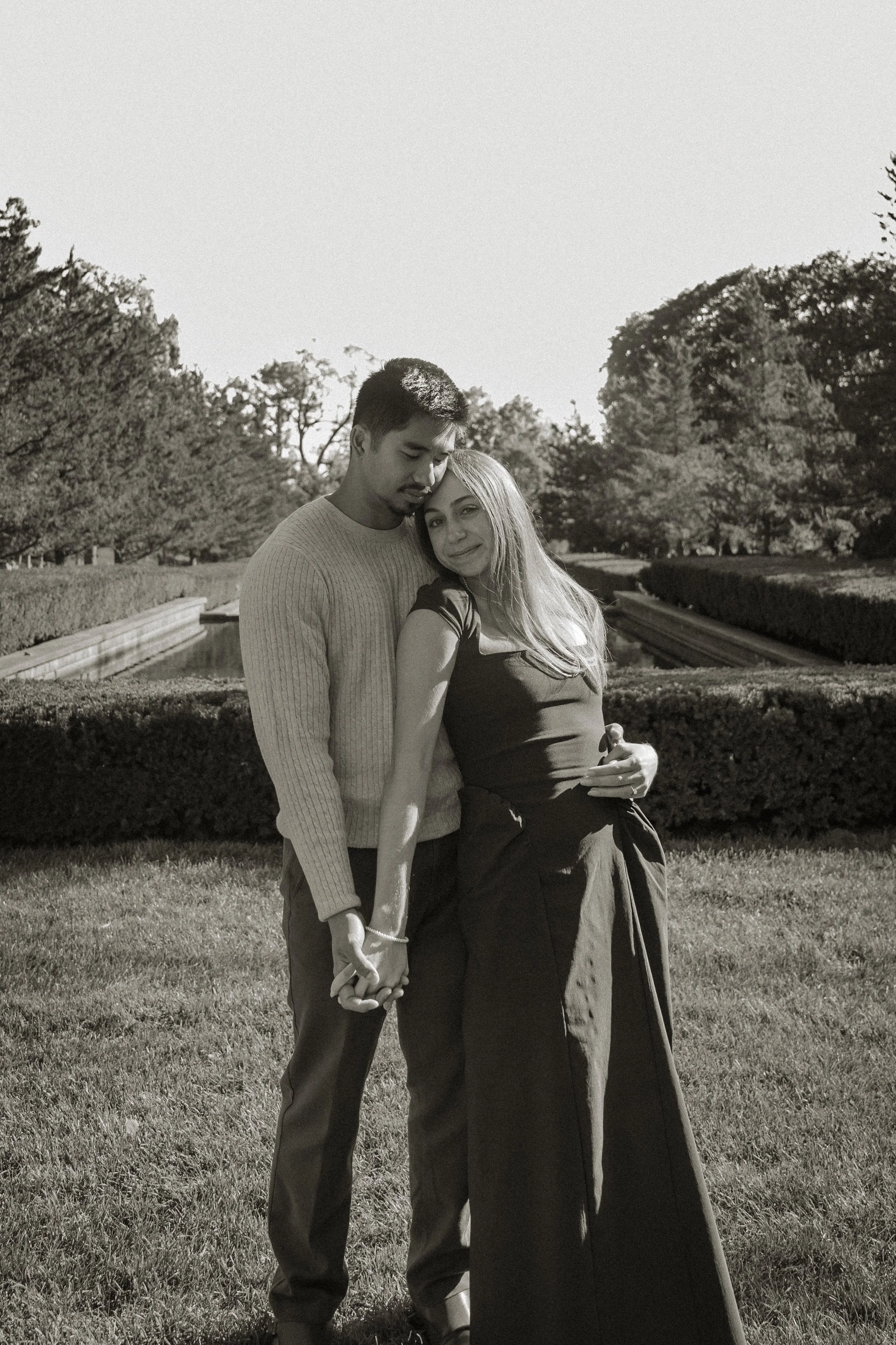A couple holding hands stands on a grassy area near a canal, with trees in the background, captured in black and white.
