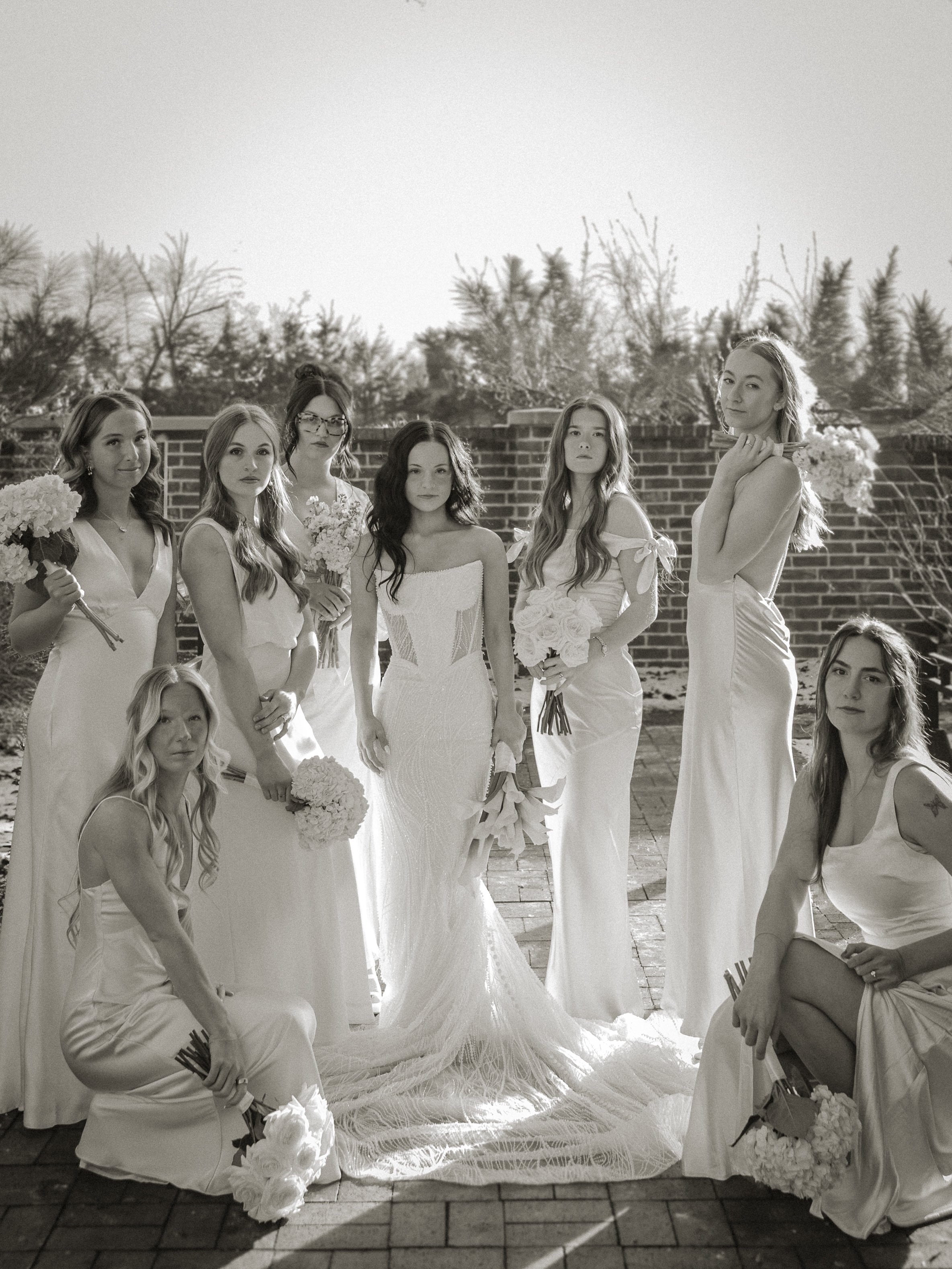 A group of nine women dressed in white wedding gowns and holding bouquets standing outdoors on a brick surface, with trees and a brick wall in the background.
