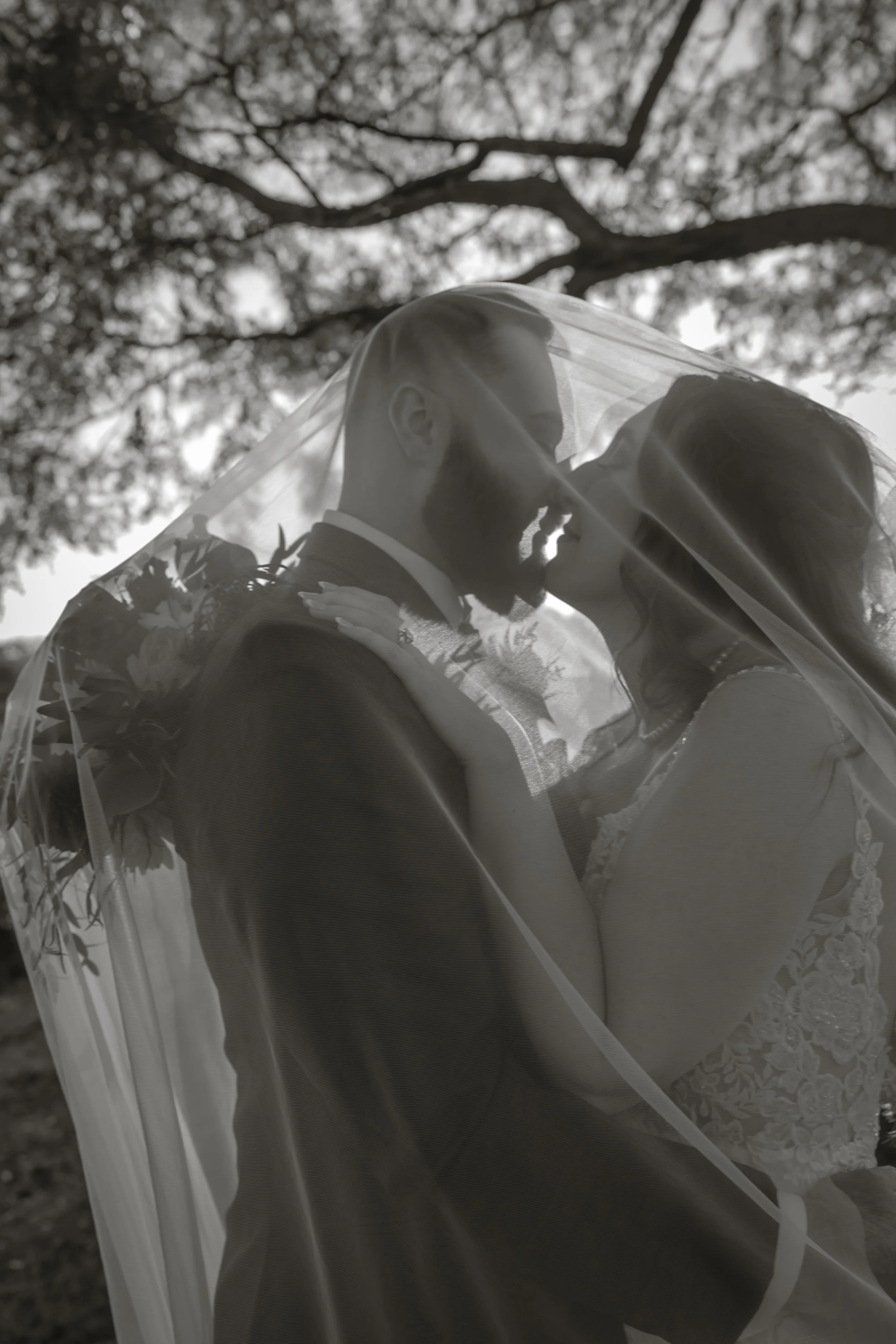 Black and white photo of a bride and groom kissing underneath a veil outdoors, with trees in the background.
