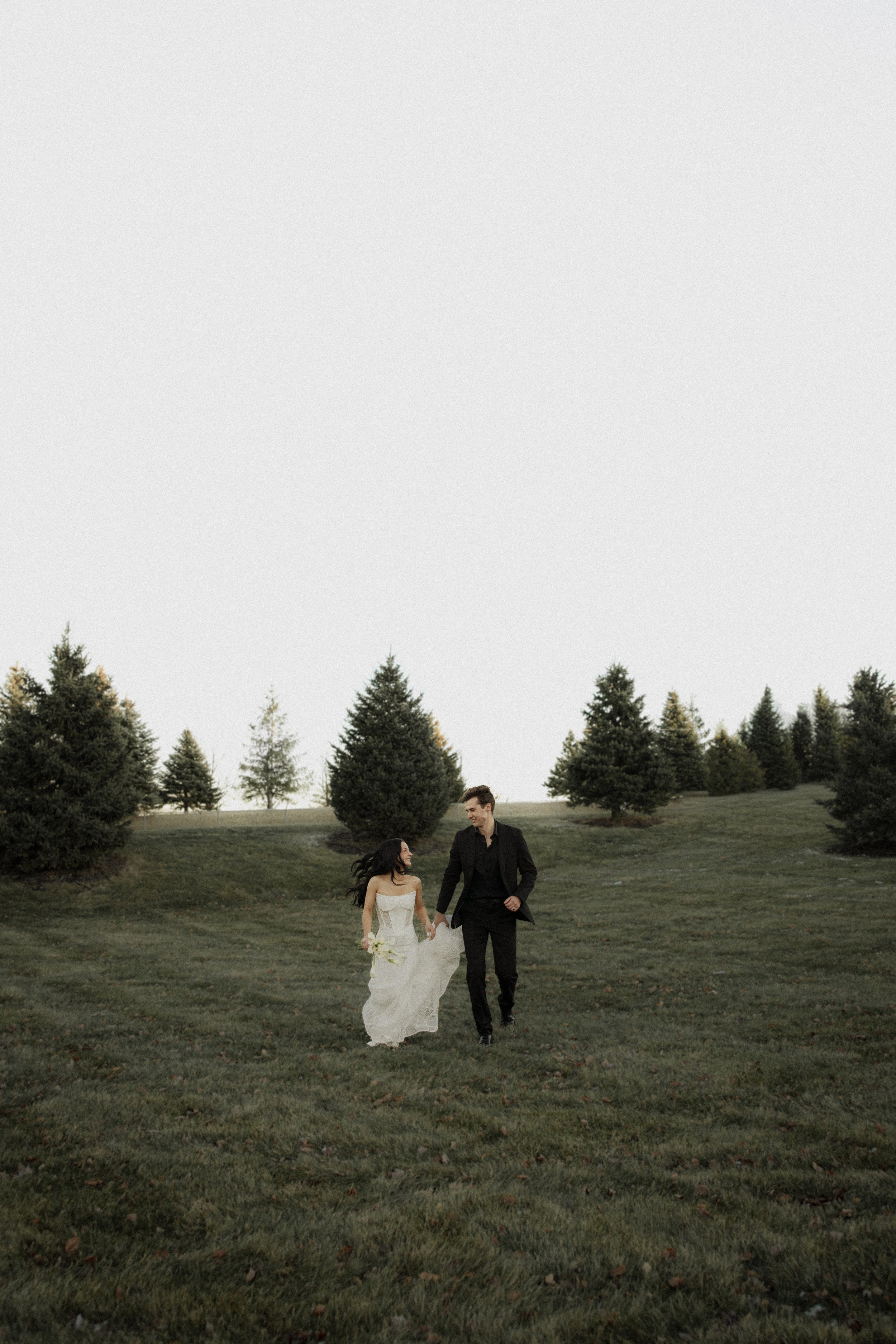 A bride and groom walking hand in hand on the grass in a park with evergreen trees in the background during daytime.