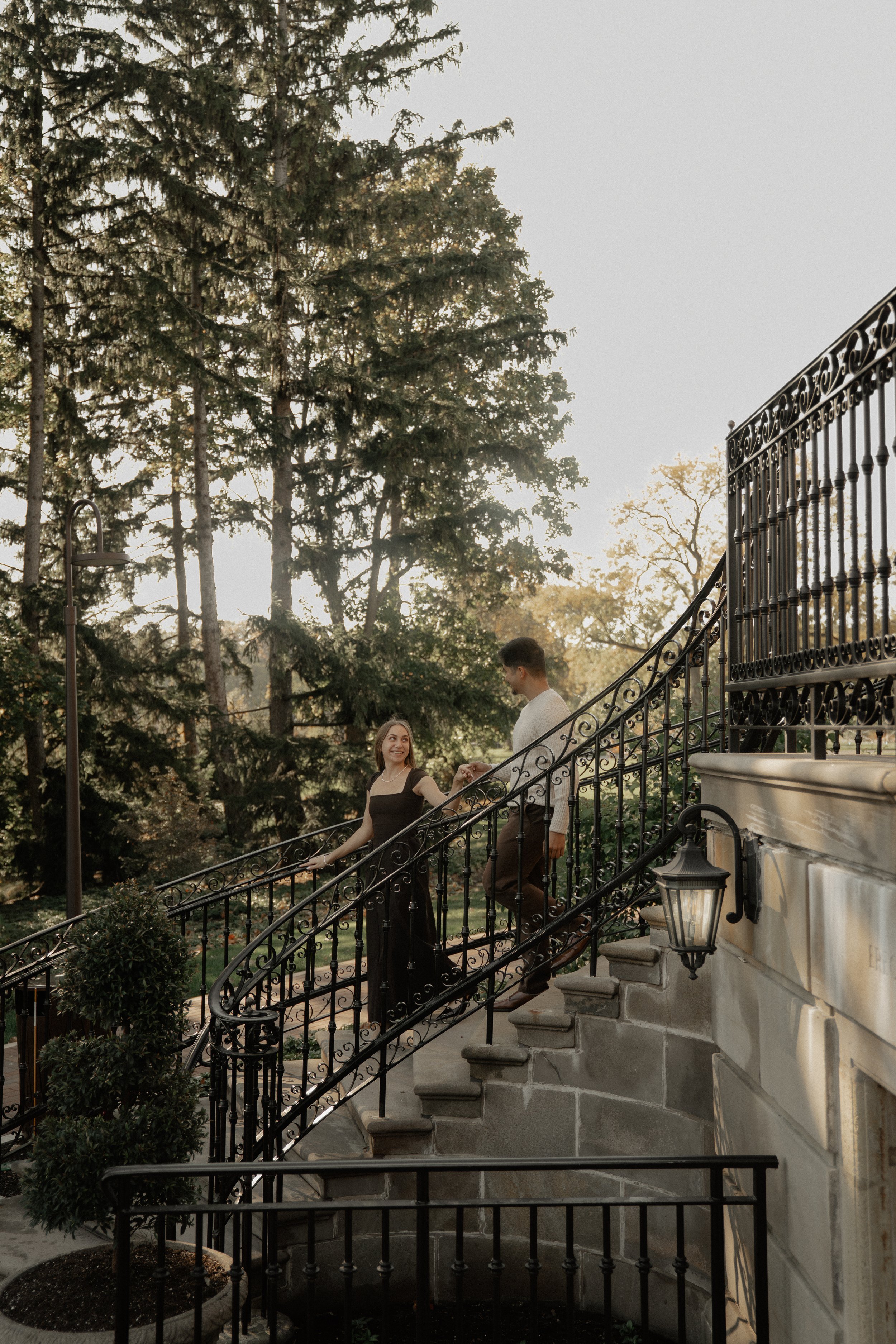 A young couple holding hands and smiling while walking up an outdoor staircase with black wrought iron railings, surrounded by trees and greenery, during late afternoon or early evening.