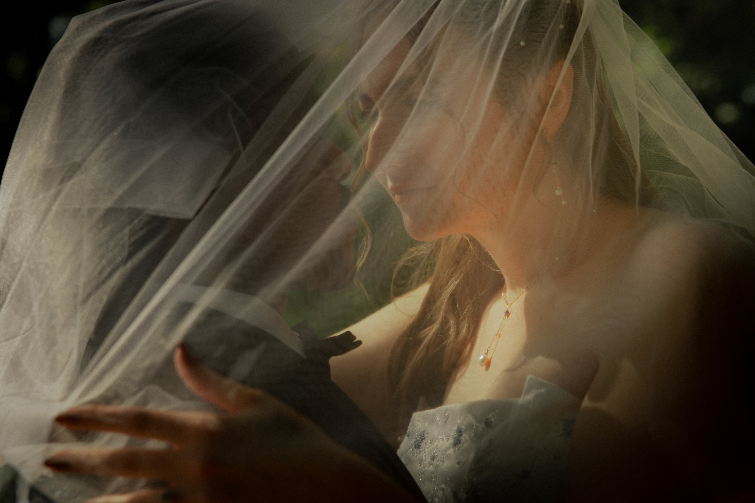 A bride and groom close together under a veil, about to kiss, with an outdoor background.