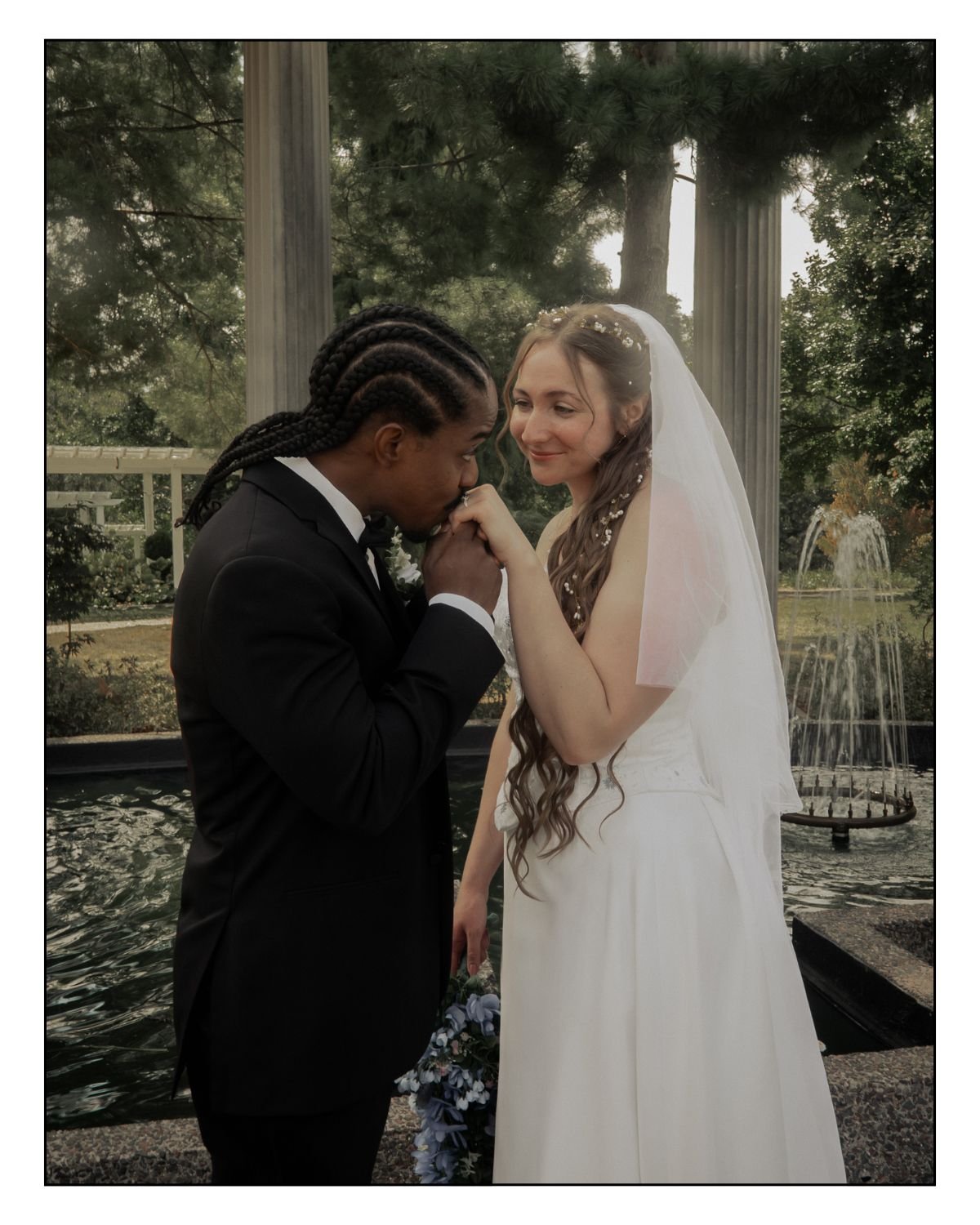 A bride and groom sharing a tender moment, holding hands, in front of a fountain and classical columns outdoors during their wedding.