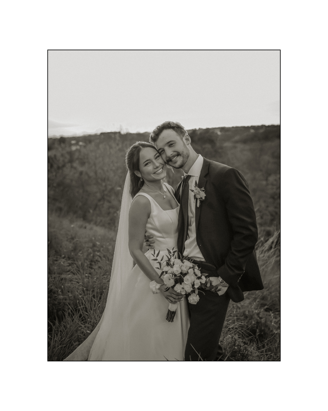 A newlywed couple, the bride in a white wedding dress holding a bouquet of flowers, and the groom in a dark tuxedo, smiling and embracing outdoors.