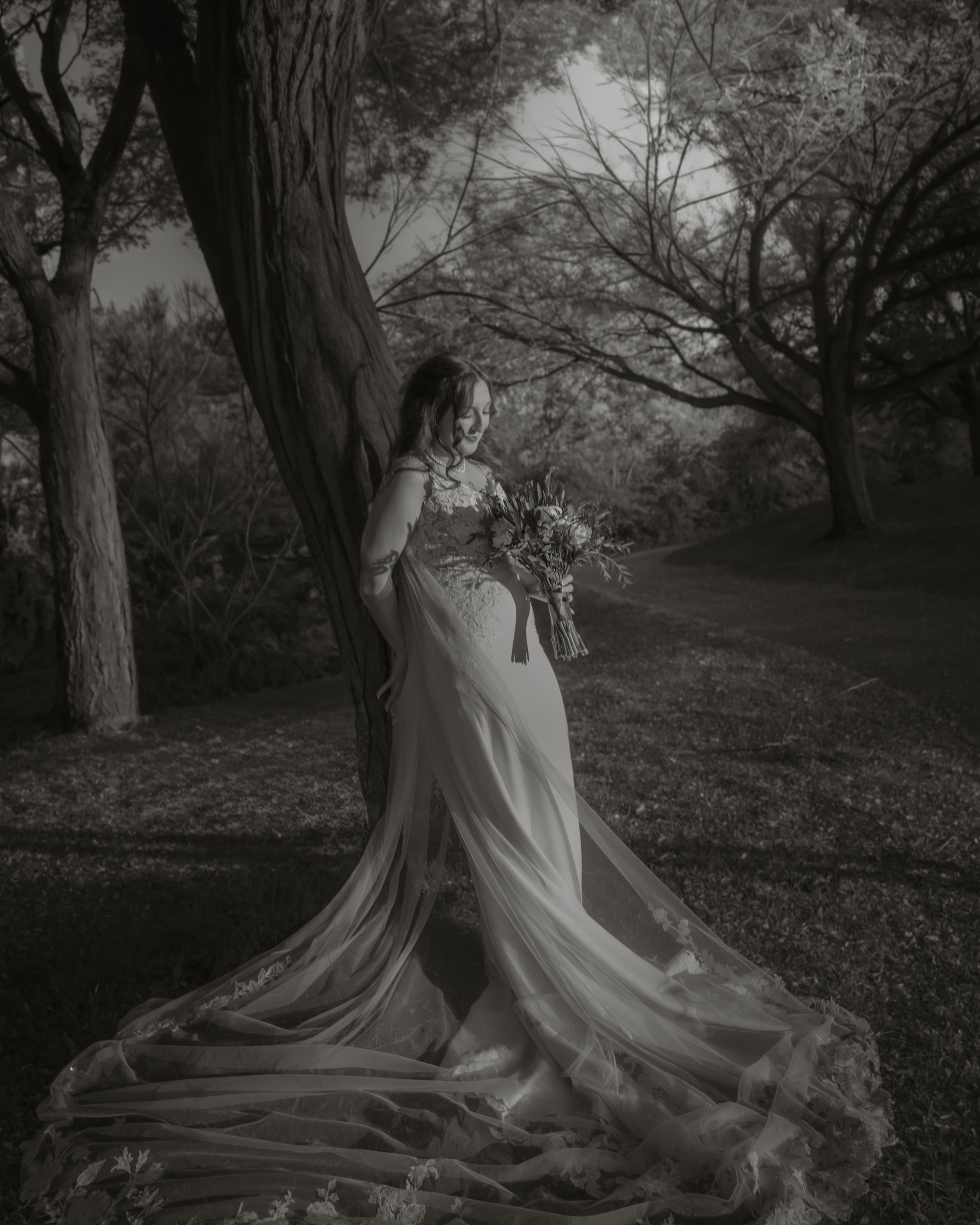A bride in a wedding gown holding a bouquet of flowers, standing outdoors next to a tree in a park or garden.