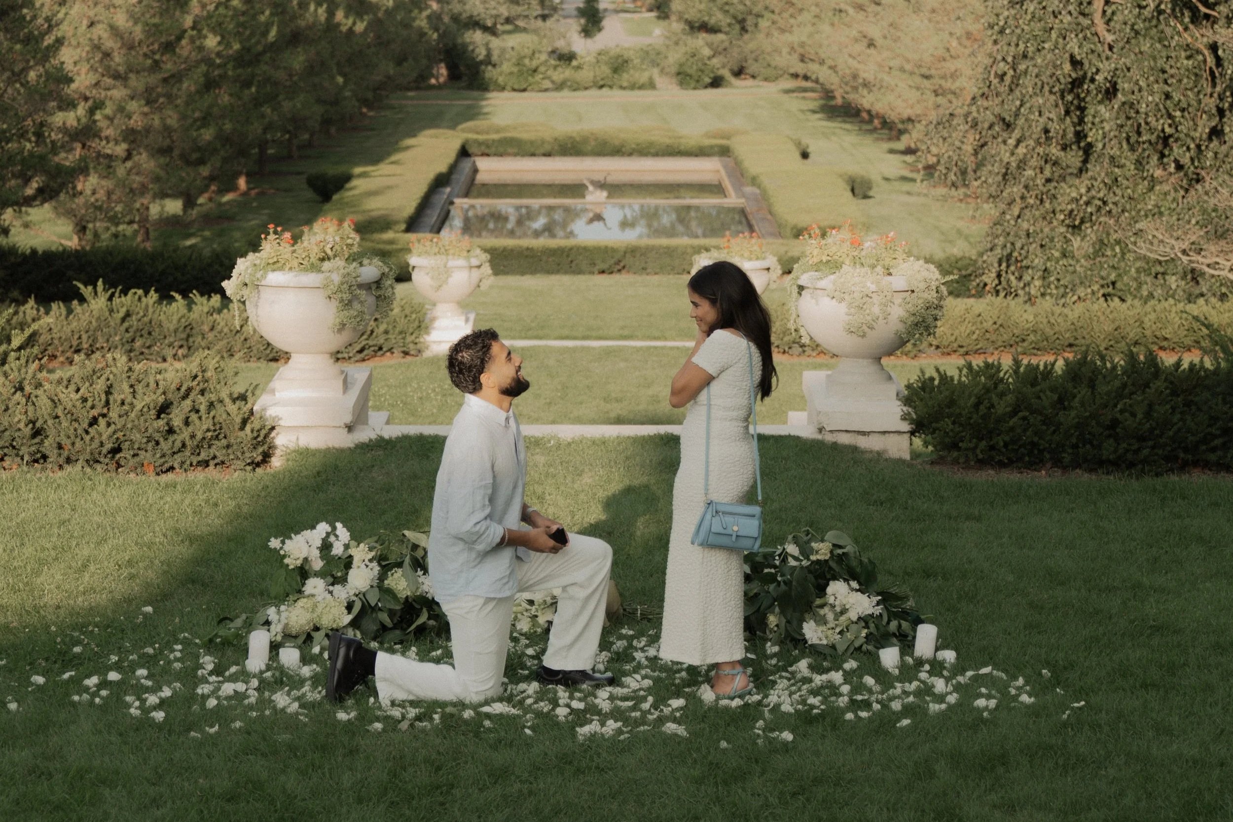 A man kneeling on one knee in snow proposes to a woman, who covers her mouth in surprise, with trees and a sunset in the background.