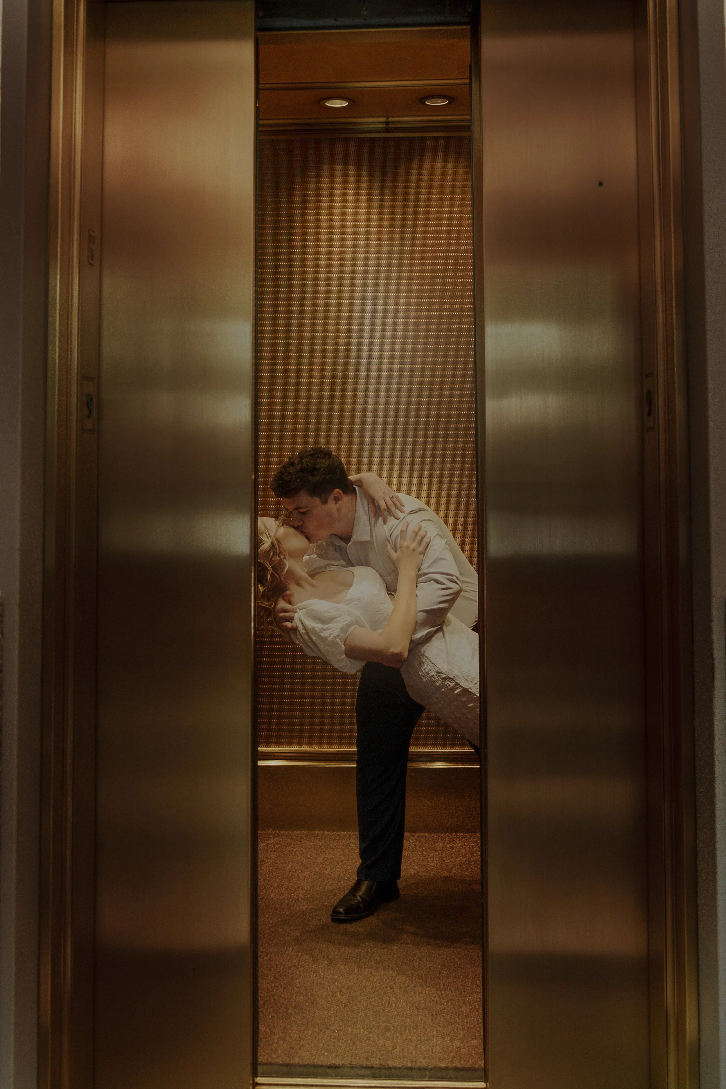 A man in dress shirt and pants is holding a woman in a white dress in a dip, sharing a kiss inside an elevator with wooden and metallic interior.