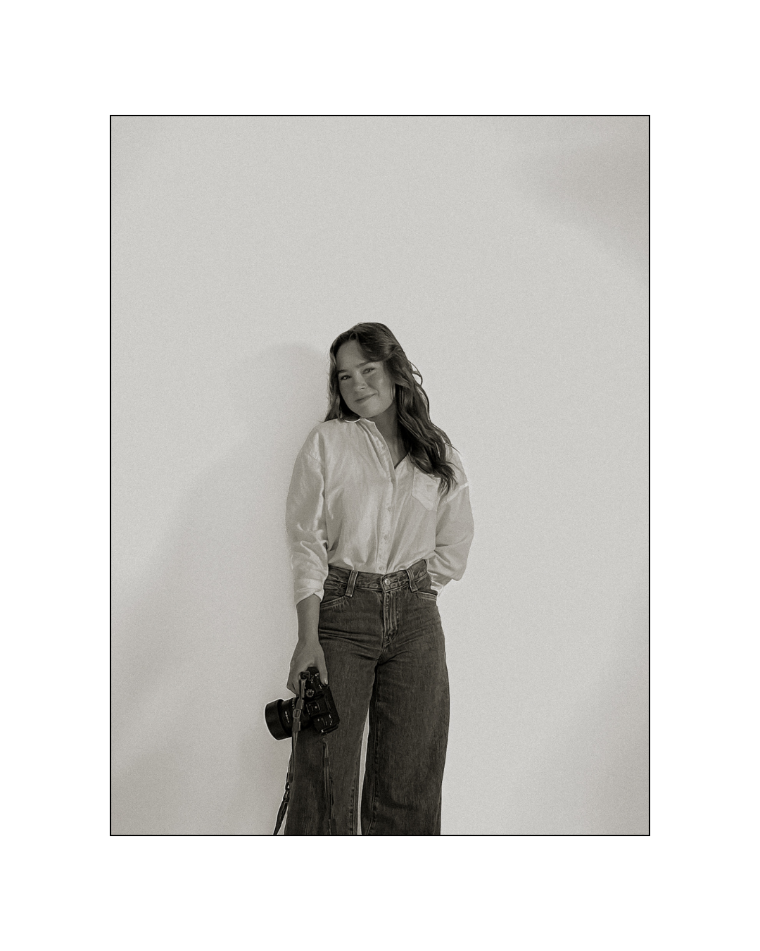 Black and white photo of a woman standing against a plain background, holding a camera in her right hand, smiling softly, with long wavy hair, wearing a button-up shirt and jeans.