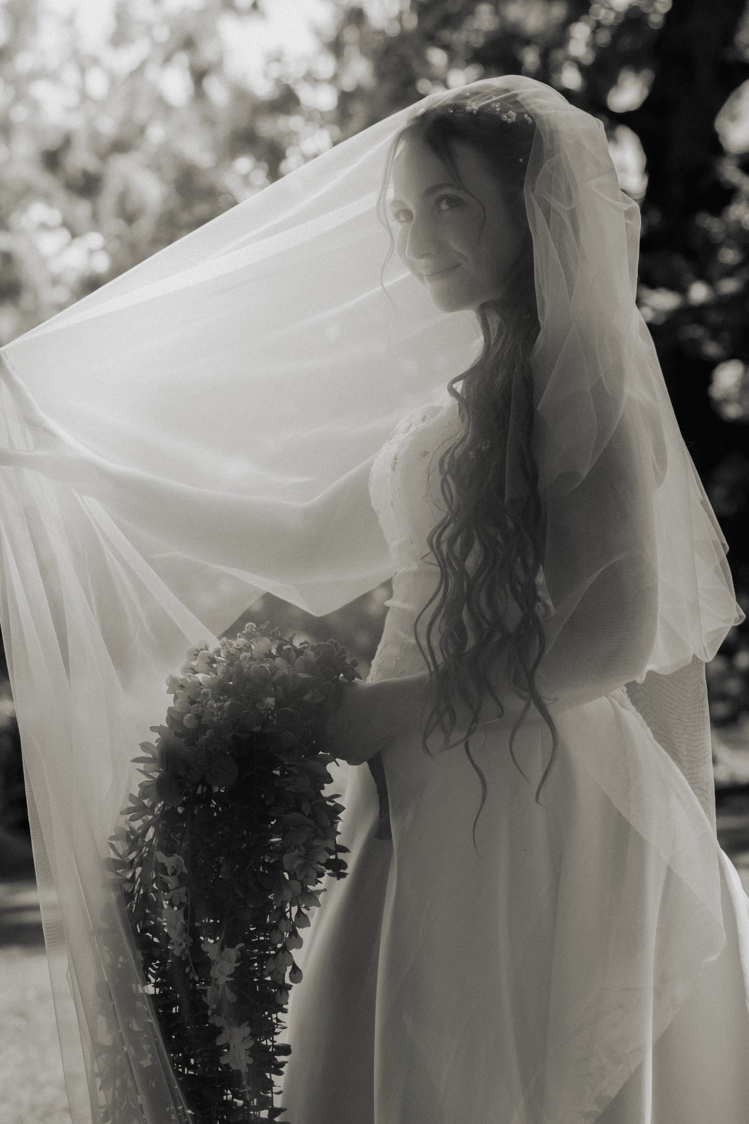 A bride holding a bouquet, wearing a wedding dress and a veil, with trees in the background.