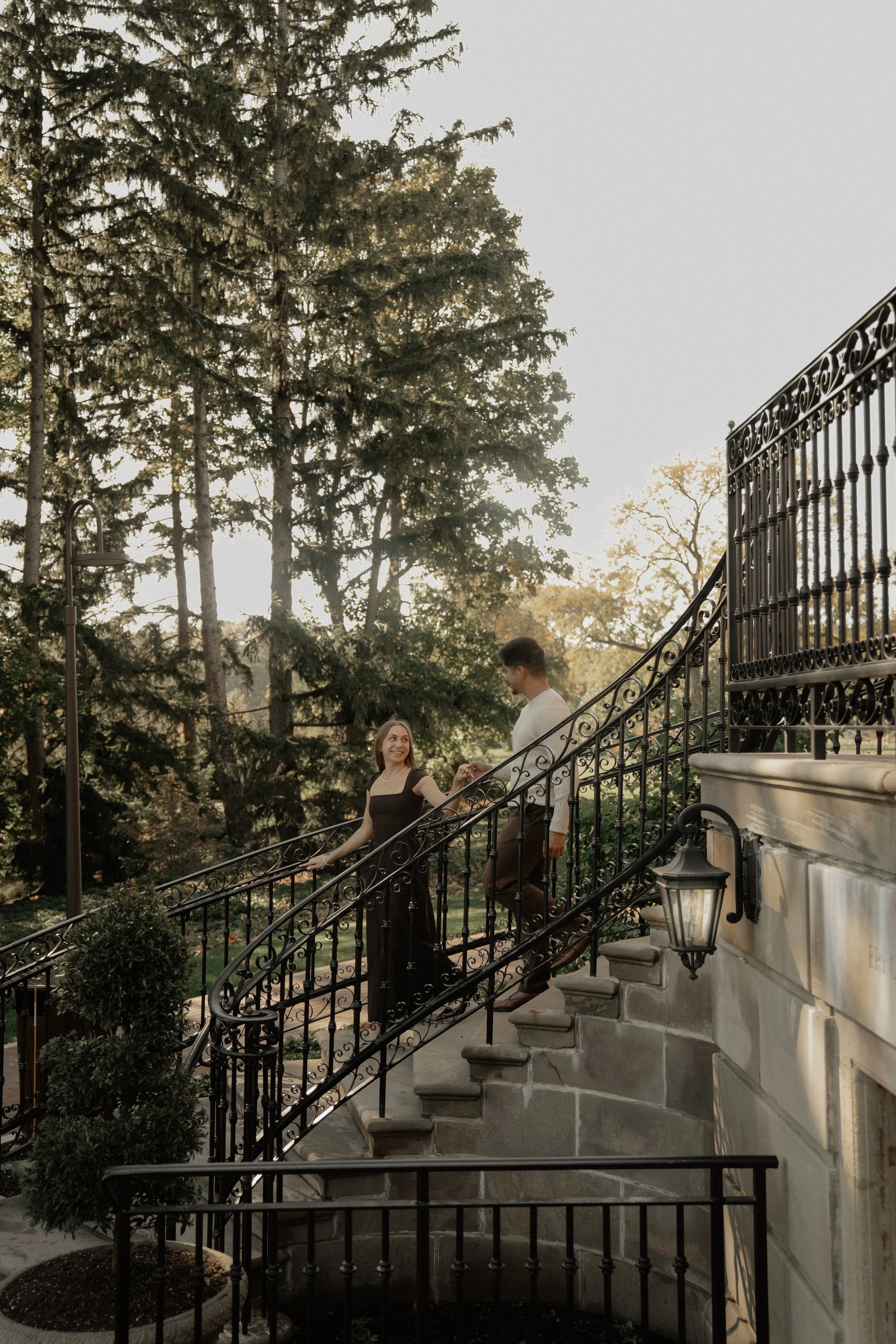 A woman and a man on a staircase outdoors, holding hands, with trees and sunlight in the background.