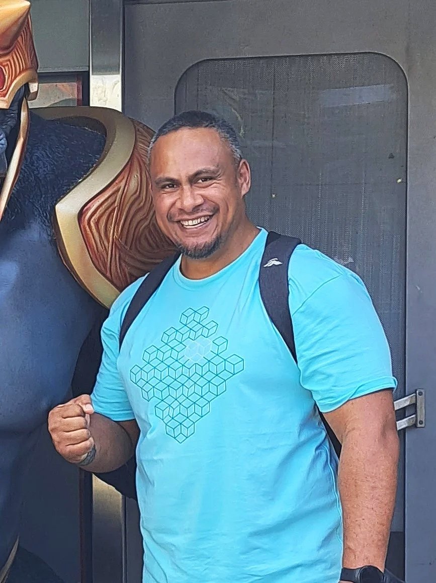 Smiling man with short hair and a beard wearing a light blue t-shirt and backpack, standing in front of a decorative horse sculpture.