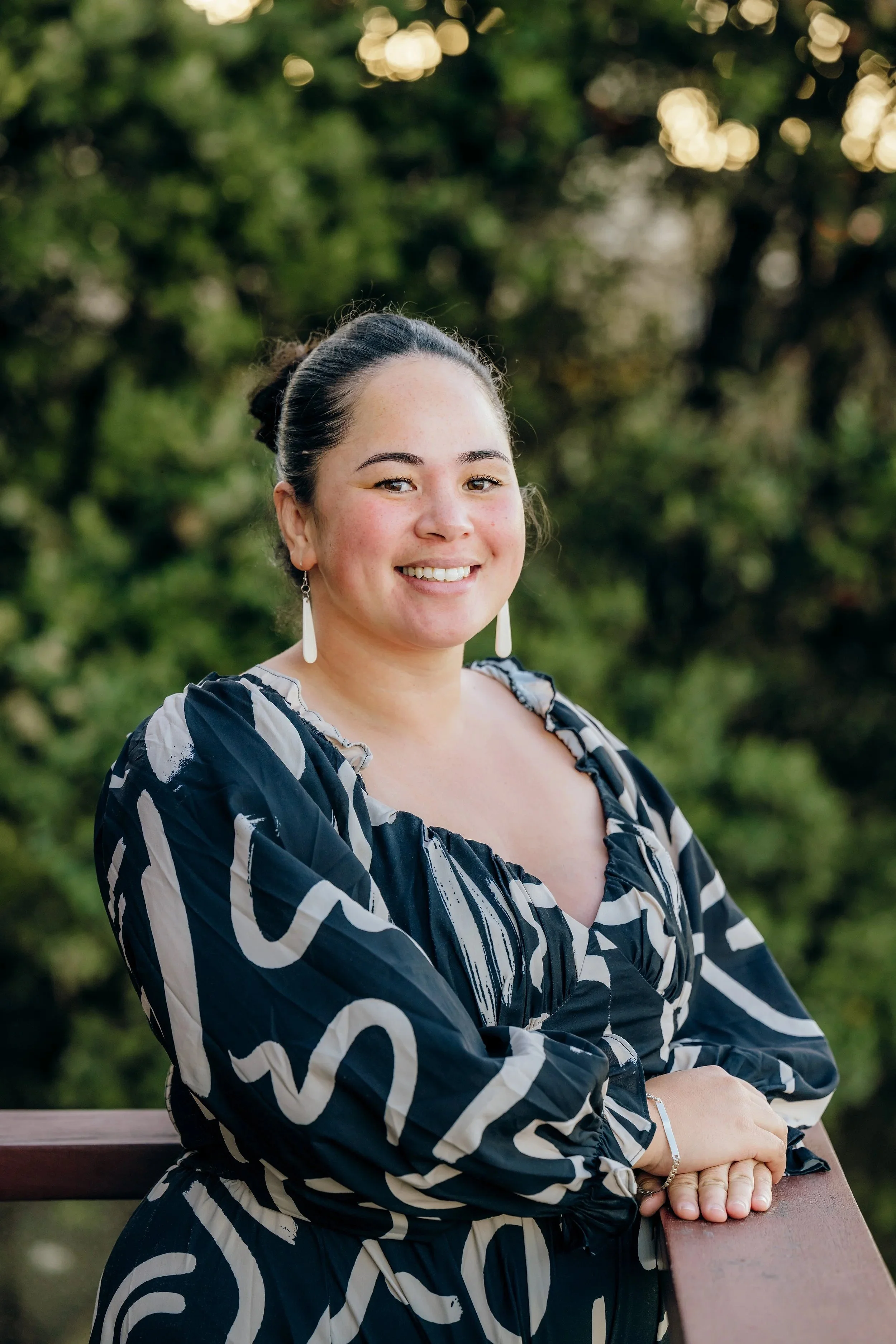 A woman with dark hair in a bun, wearing a black and white patterned blouse and white earrings, smiling while leaning on a wooden railing outdoors. Blurred green trees and warm light bokeh are in the background.