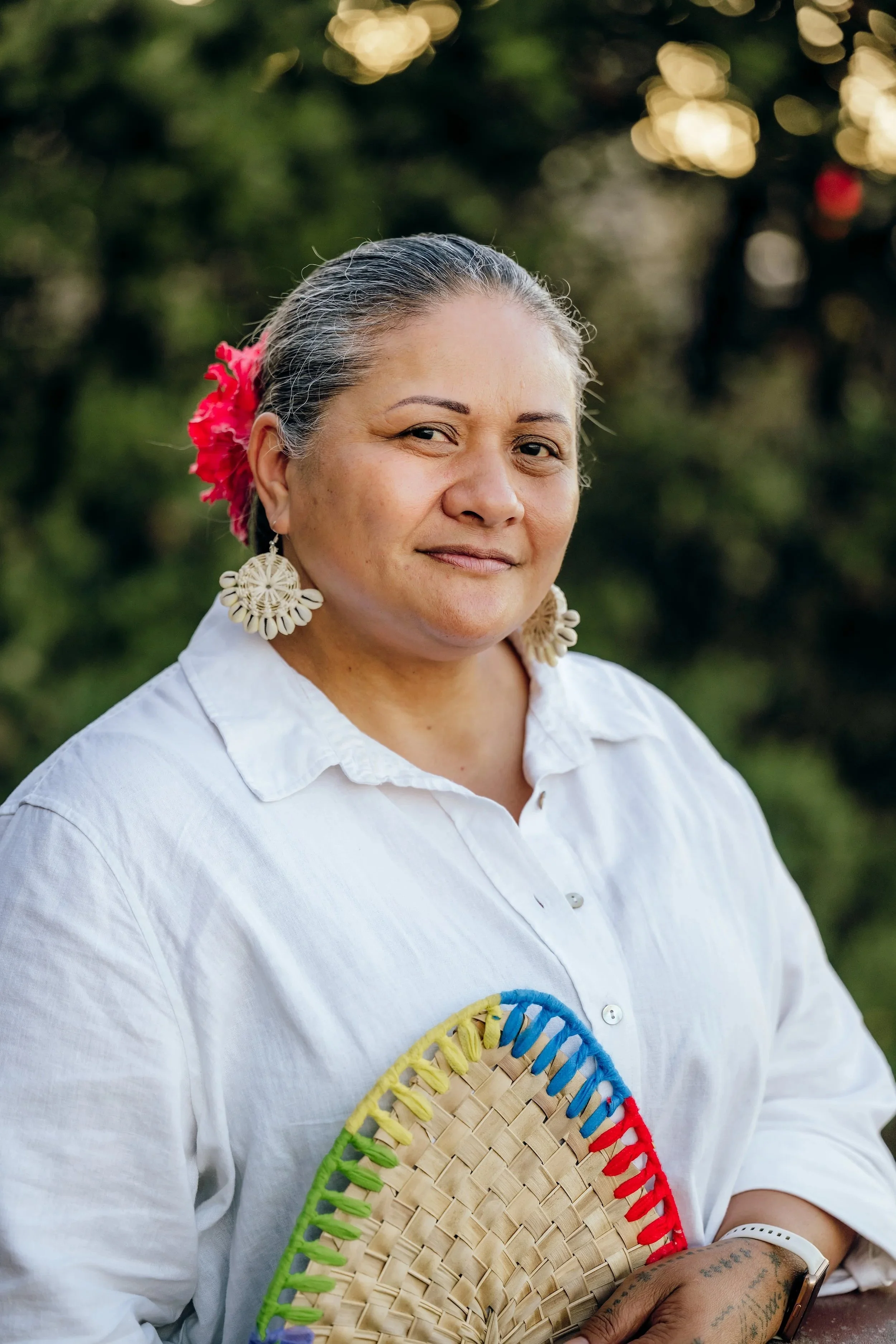 A woman with gray hair tied back, wearing a white shirt, large woven earrings, and a pink flower in her hair, holding a colorful woven fan, outdoors with blurred greenery and lights in the background.