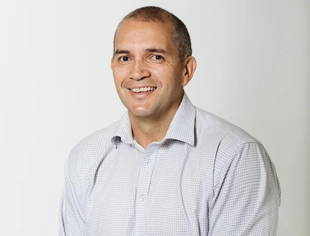 A smiling man in a light-colored button-up shirt posing against a plain white background.