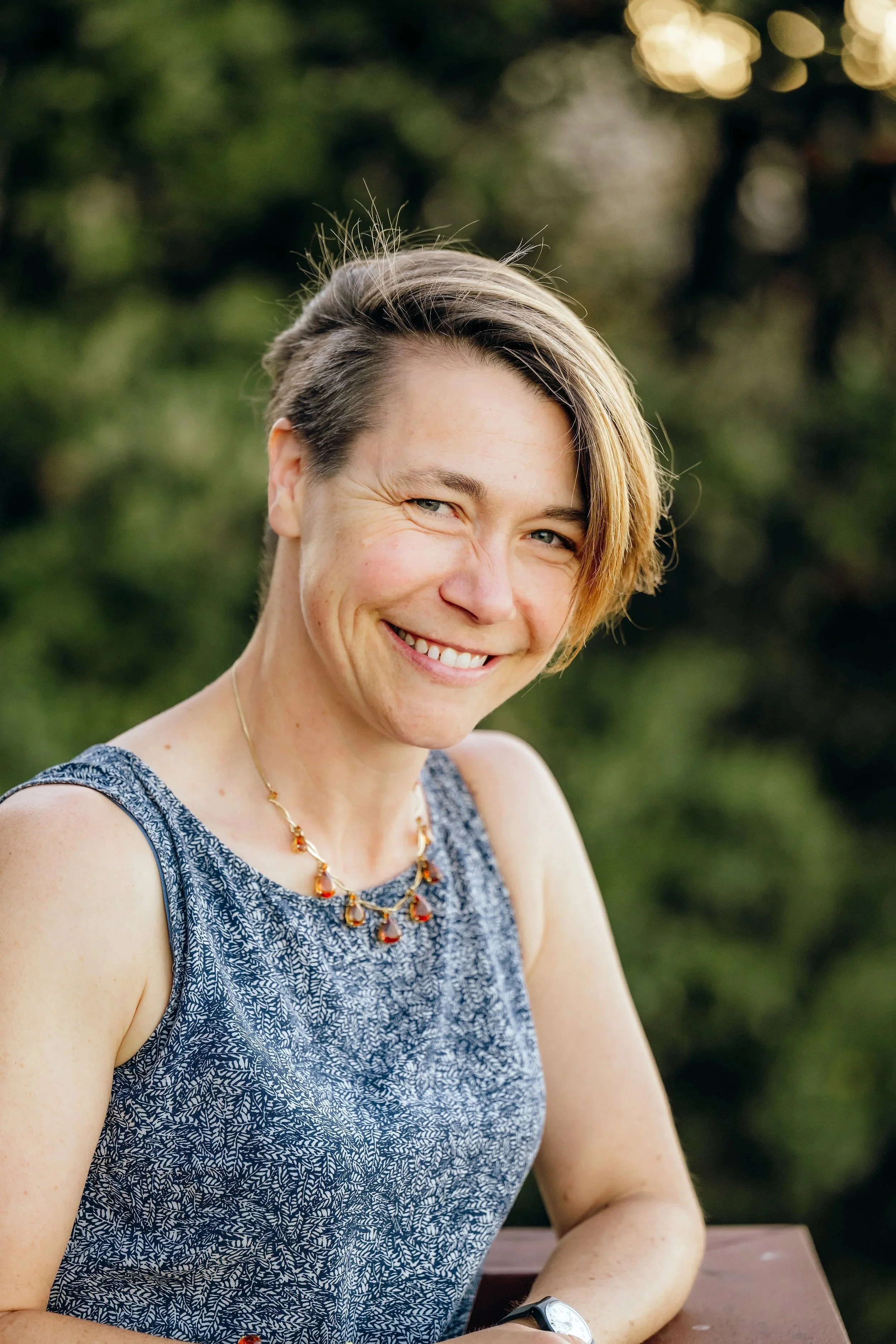 A smiling woman with short hair, wearing a patterned sleeveless dress, a necklace, and a watch, sitting outdoors at a wooden table with a blurred green background.