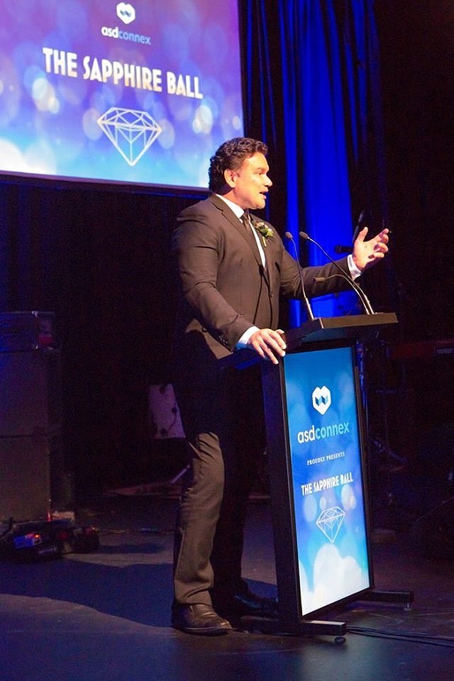 A man in a dark suit and bow tie standing at a podium with a microphone, holding a tablet, speaking at an event called The Sapphire Ball organized by asdconnex, with a large screen behind him displaying the event's logo and name.