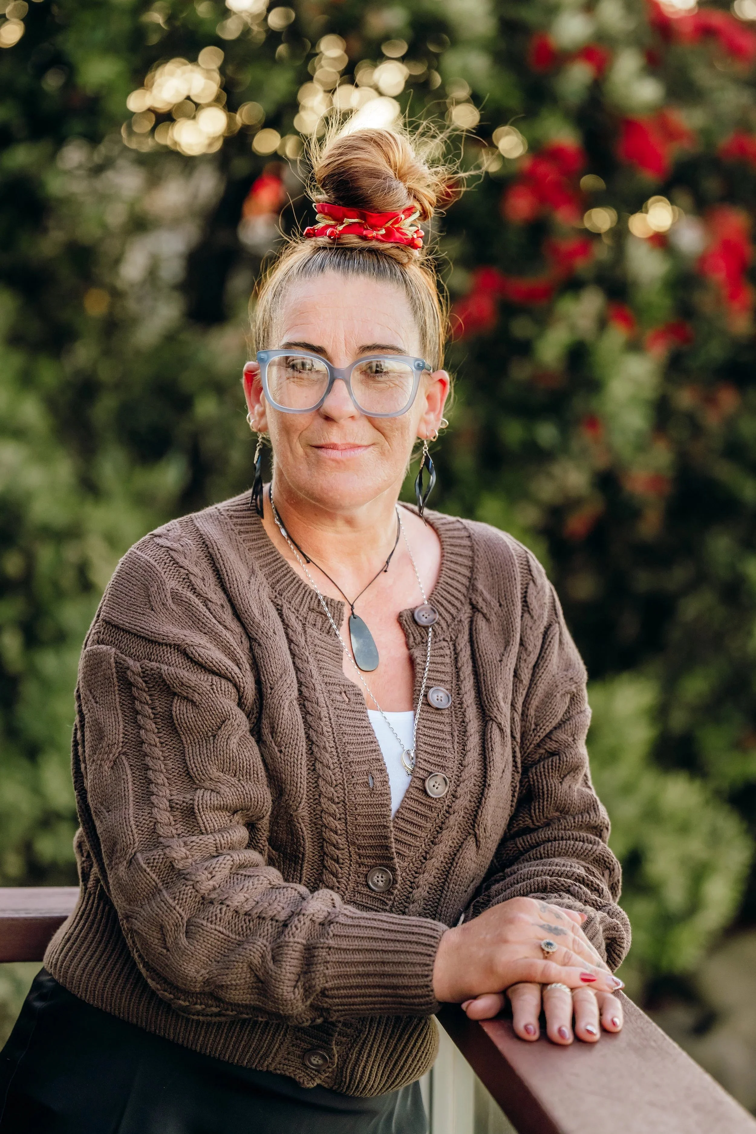 A woman with glasses and earrings, wearing a brown cable-knit cardigan and a white top, sitting outdoors on a wooden bench with her hands resting on the rail. She has her hair tied up in a high bun with a red scrunchie. The background shows blurred trees with red and green foliage.