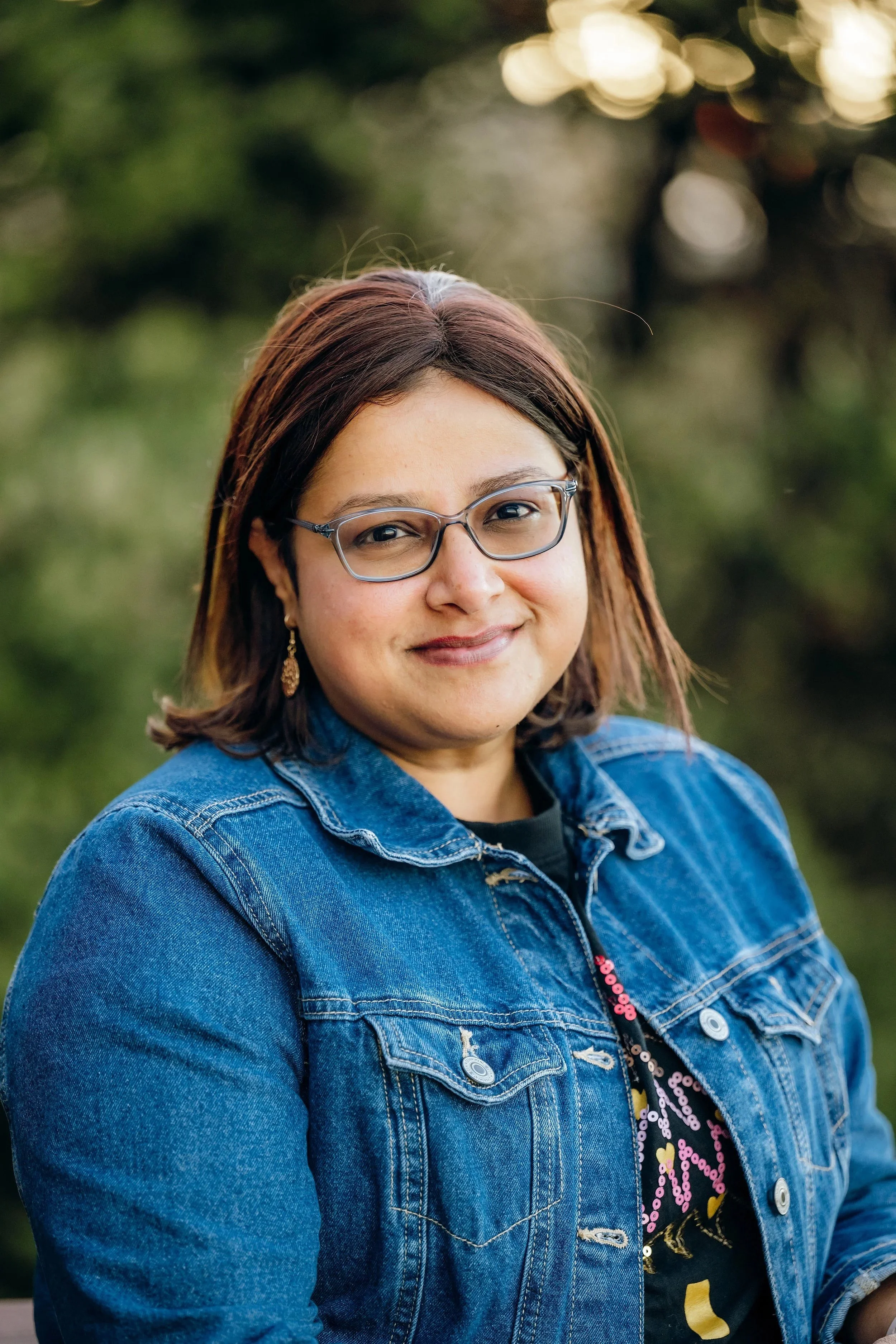 A woman with shoulder-length brown hair and glasses, wearing a denim jacket and a black top with colorful patterns, smiling outdoors with blurred greenery in the background.