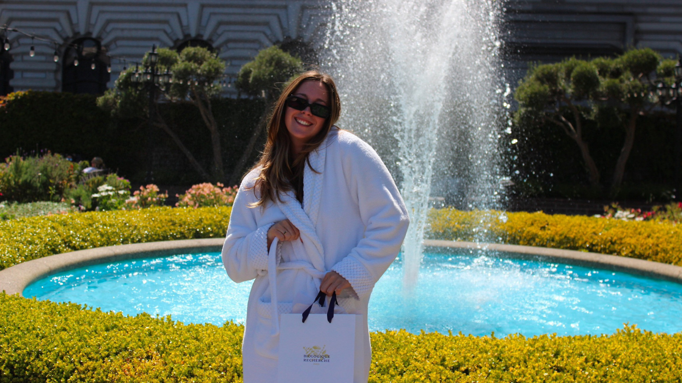 Woman holding a Biologique Recherche shopping bag after a facial, standing in a sunlit park in San Francisco.