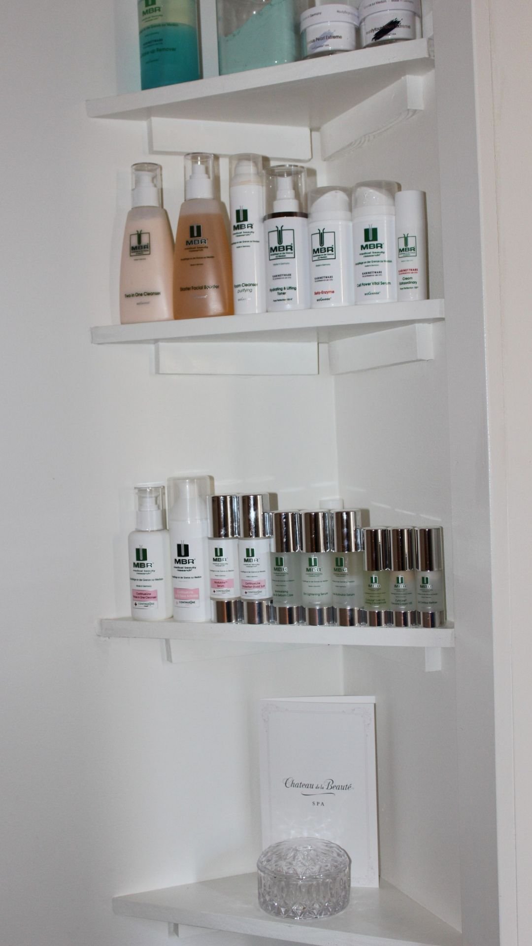 Shelves in Esthetician Aislinn’s treatment room display a curated selection of MBR Cosmetics skincare products, known for promoting radiant and revitalized skin