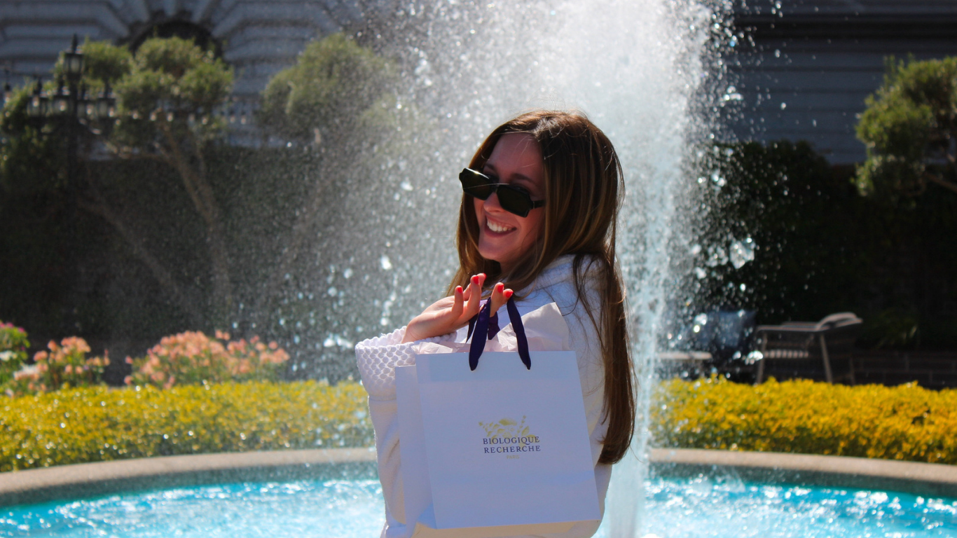 Smiling woman in sunglasses and spa robe holding a Biologique Recherche shopping bag by a sunlit fountain at Nob Hill, San Francisco