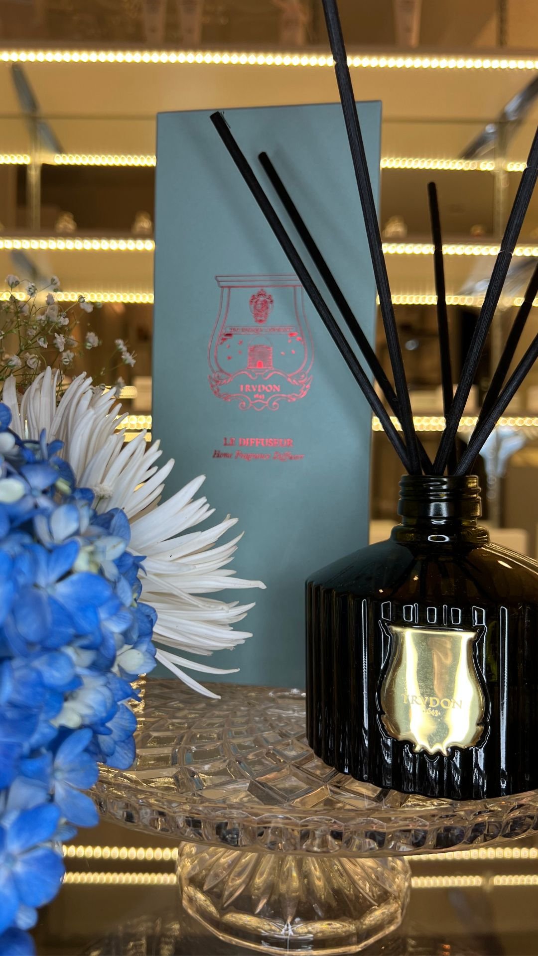 Luxury Cire Trudon reed diffuser displayed on a cut-glass pedestal with a turquoise box and delicate blue and white flowers, photographed in an elegant spa retail setting