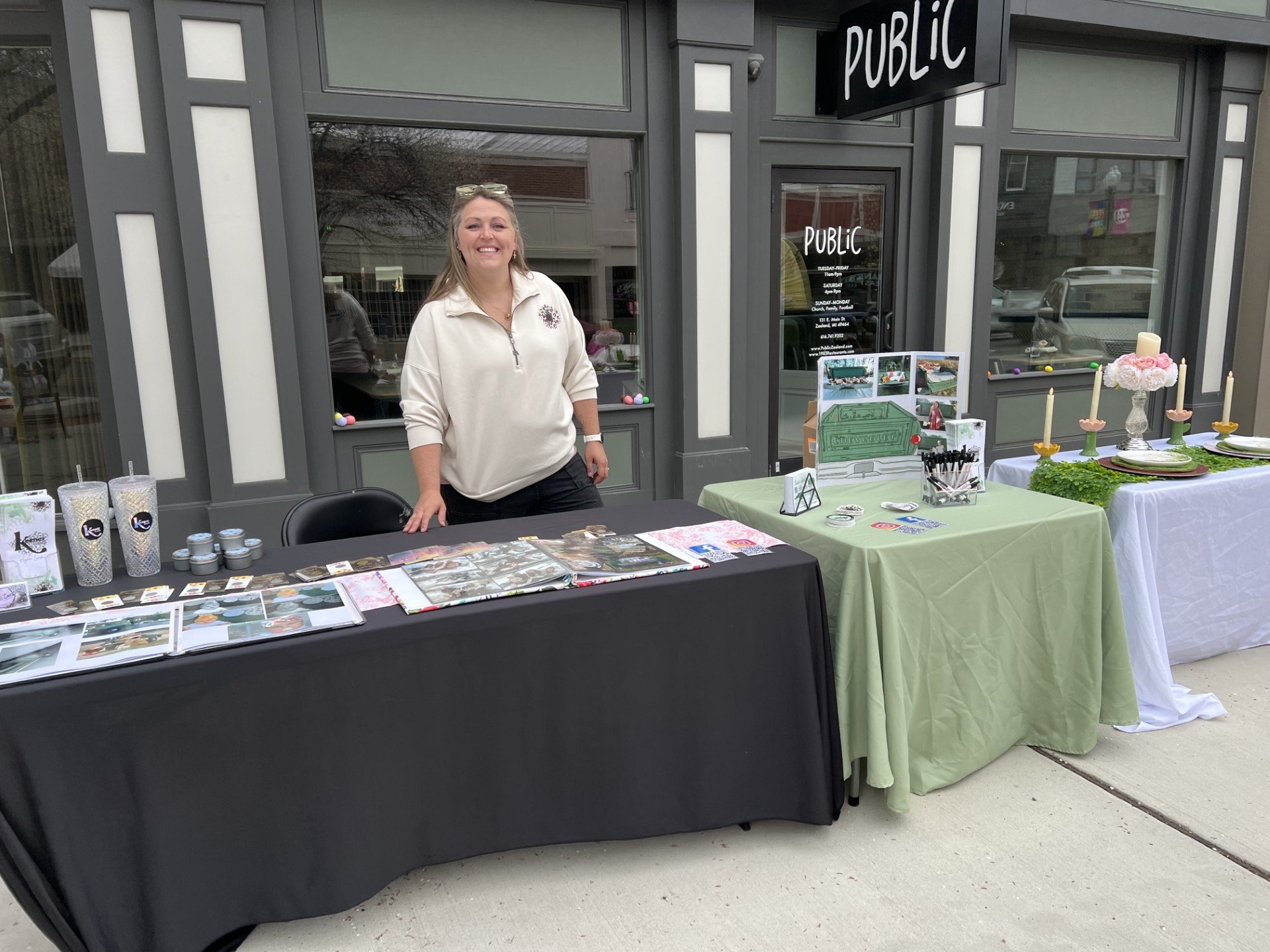 A woman stands behind a table with a black tablecloth, displaying various items such as photos, cups, and small products. Two other tables are set up nearby with a green and white tablecloth, showcasing more items and decorations outside a storefront