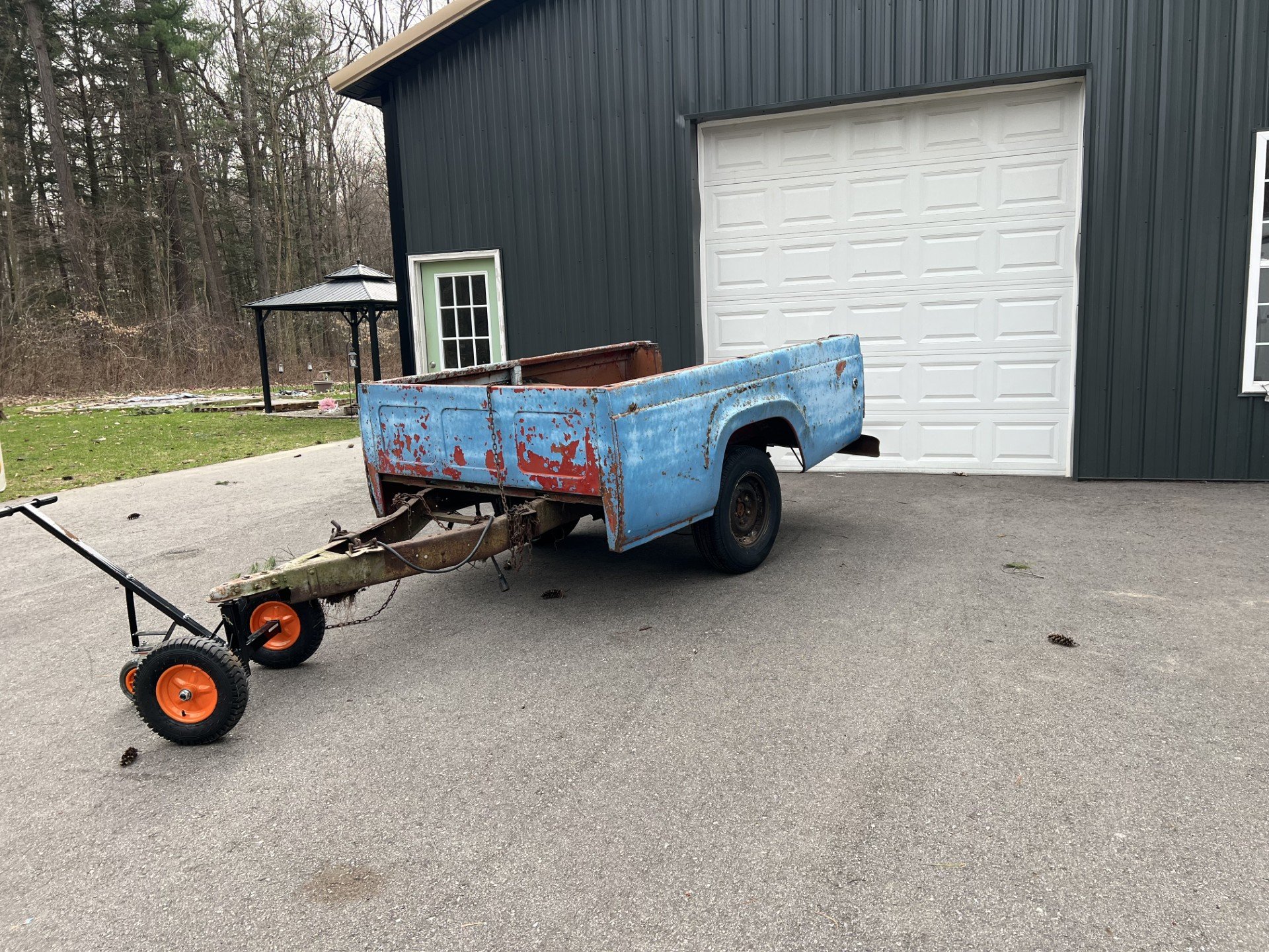 Old blue truck bed trailer parked outside a garage with a gray door and black siding. The trailer is mounted on wheels and surrounded by a concrete driveway and a grassy area with trees.