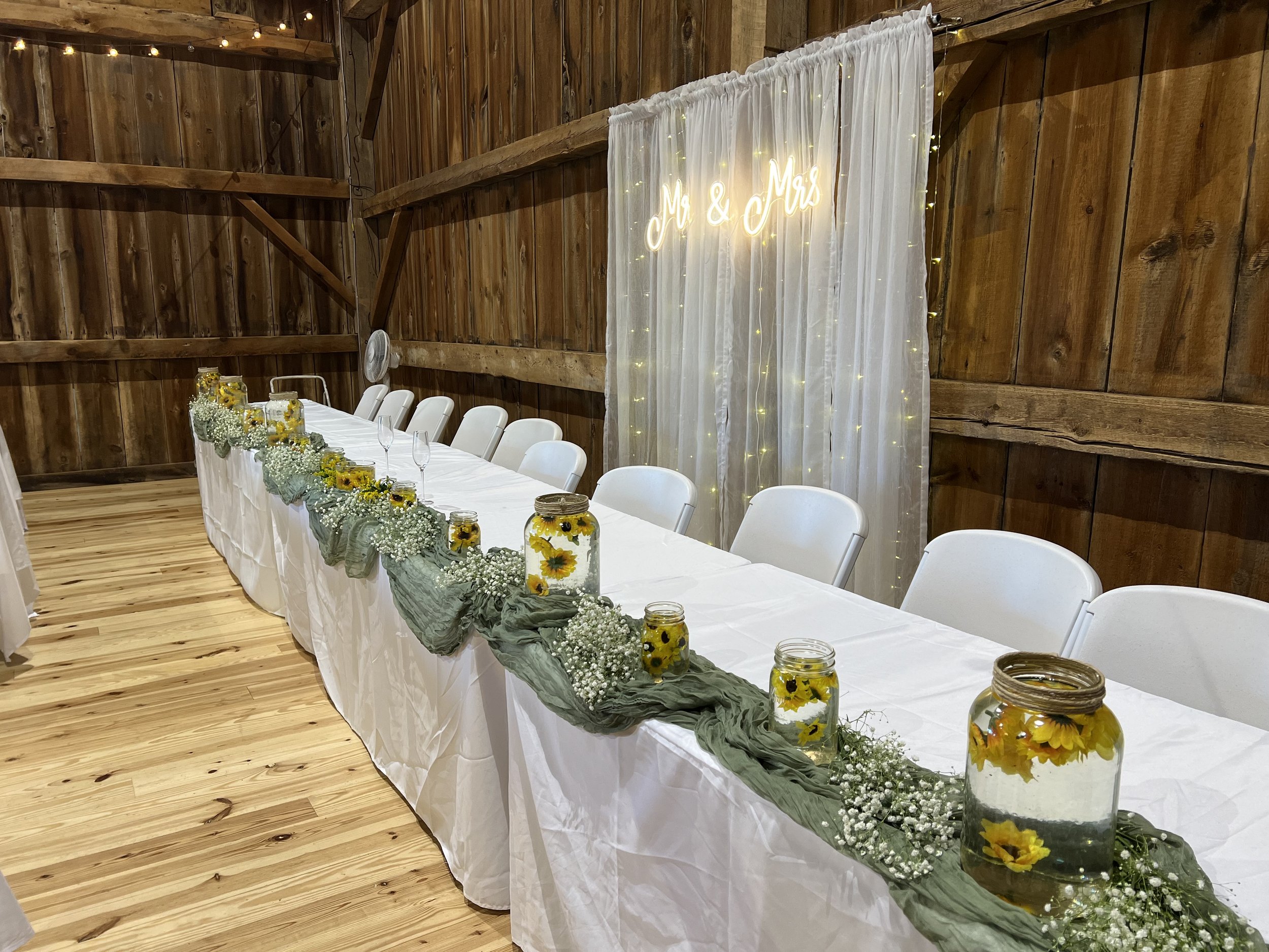 Rustic wedding reception table setup with white tablecloth, mason jars with floating flowers, greenery, and baby's breath, against a wood barn backdrop with a "Mr & Mrs" sign and string lights.
