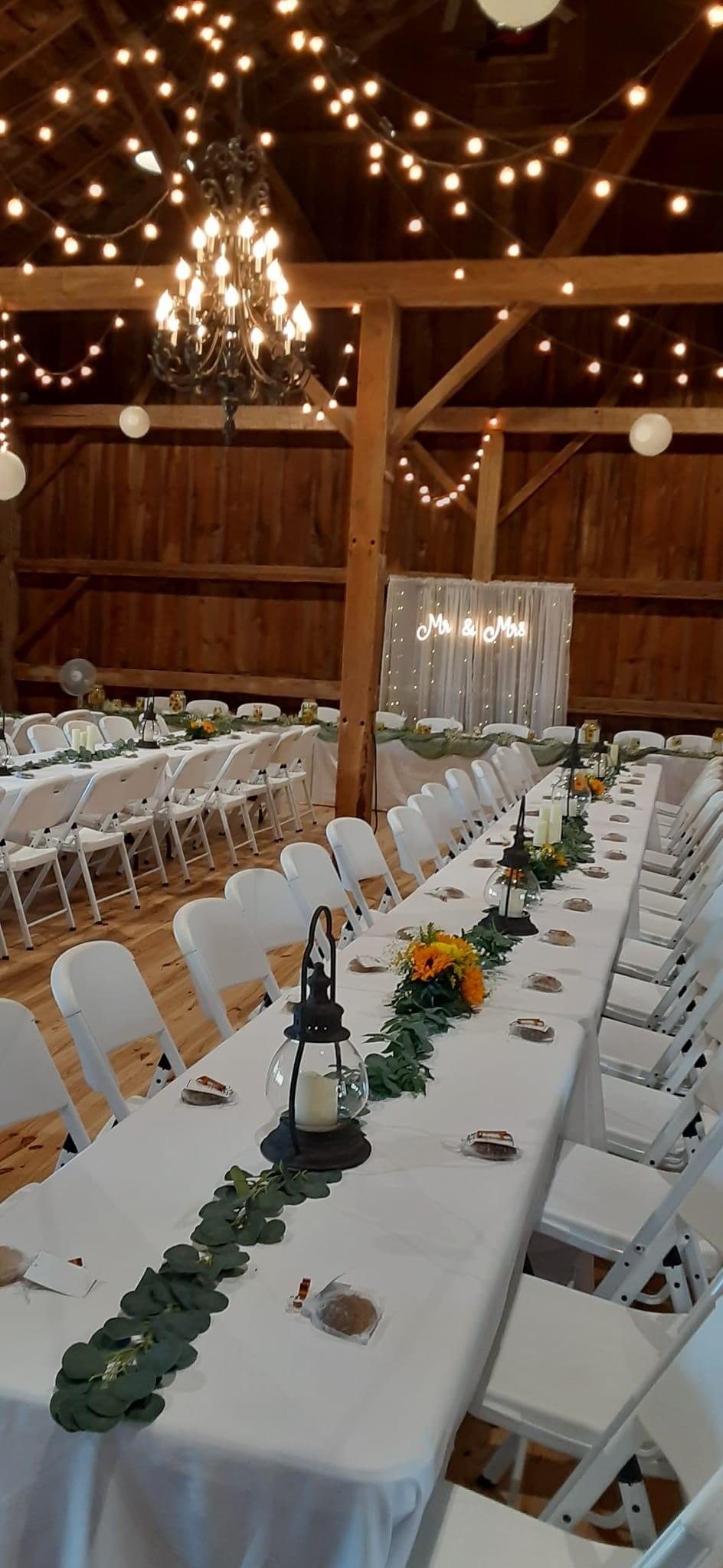 Wedding reception setup in a barn with long tables, white chairs, string lights, and rustic decorations.