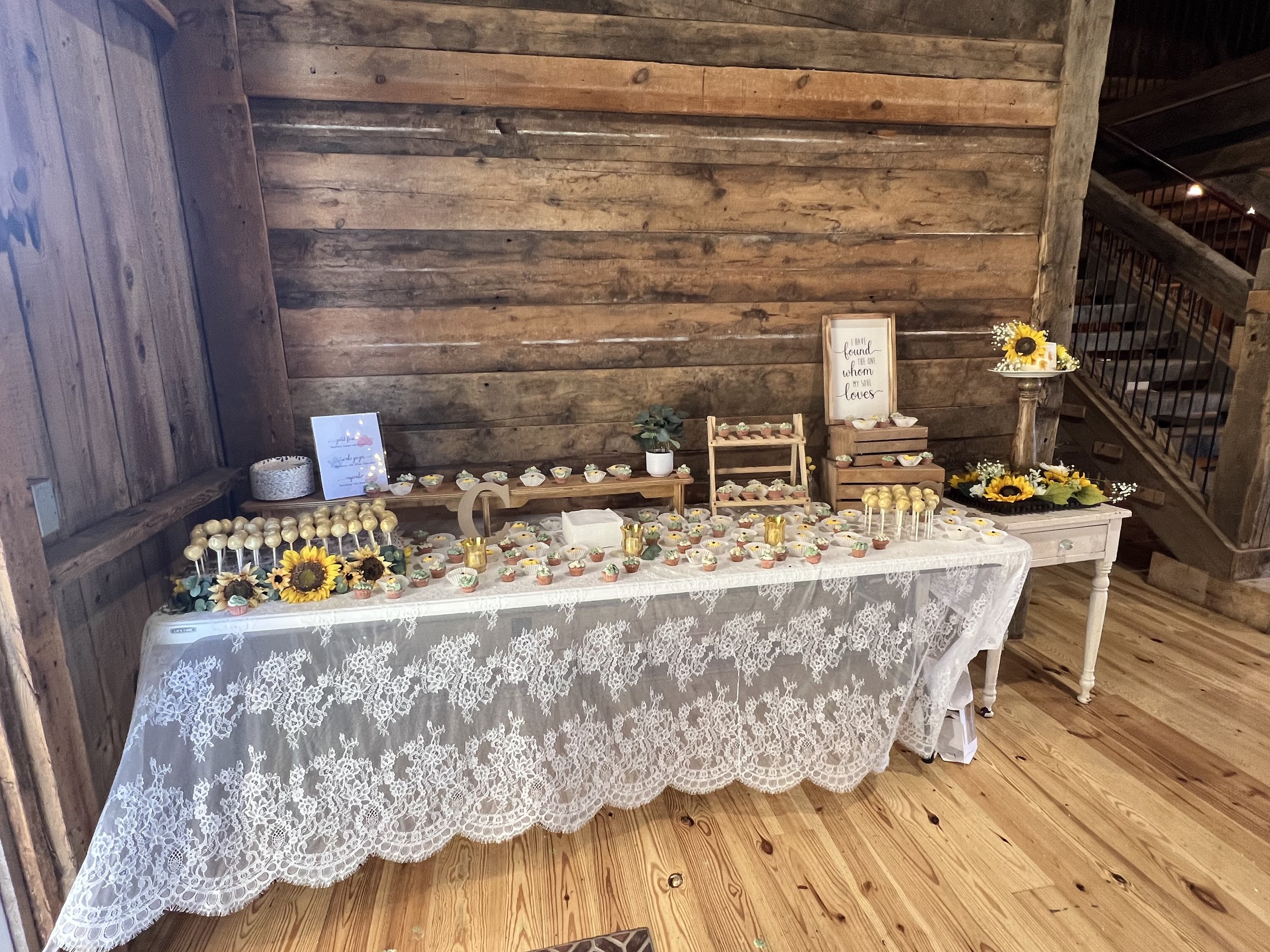 Rustic dessert table with lace tablecloth, assorted bite-sized desserts, cake pops, sunflowers, and decorative signs against a wooden wall.
