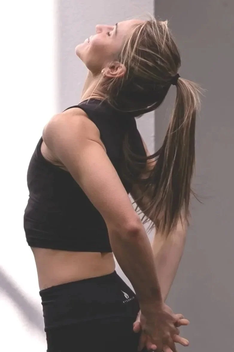 A woman with long hair tied in a ponytail stretching her back in a gym or fitness environment.