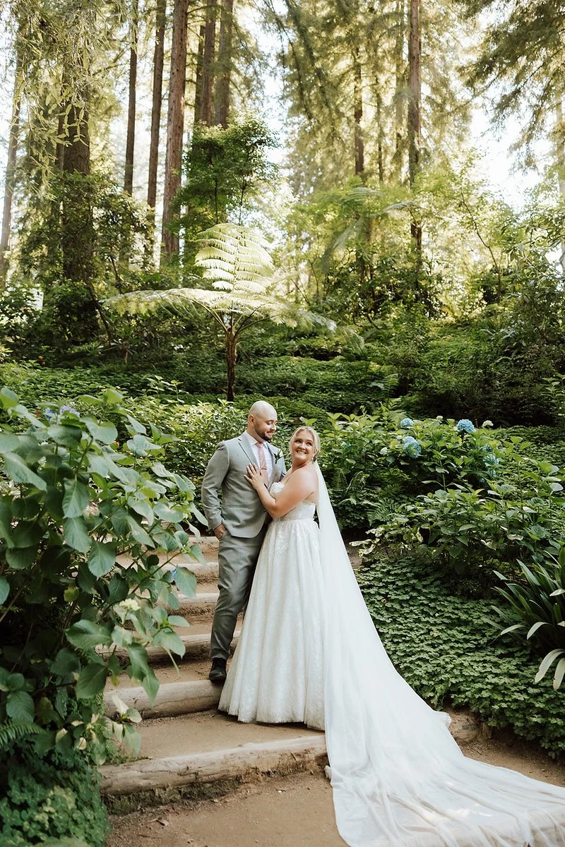 Bride and groom standing on a wooded garden path, surrounded by lush green plants and trees, with sunlight filtering through the canopy.