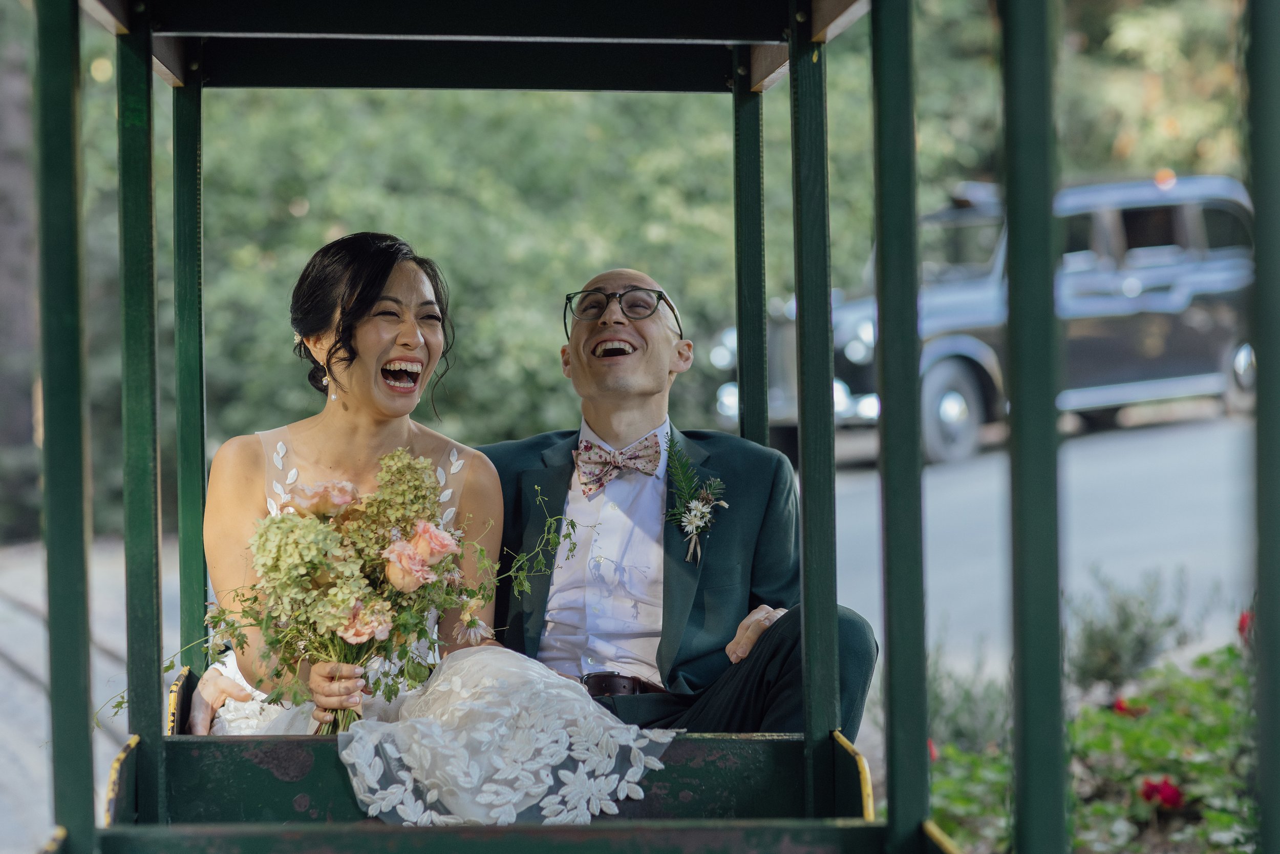A bride and groom sitting inside a carriage, laughing joyfully during their wedding, with a blurred vintage car in the background.