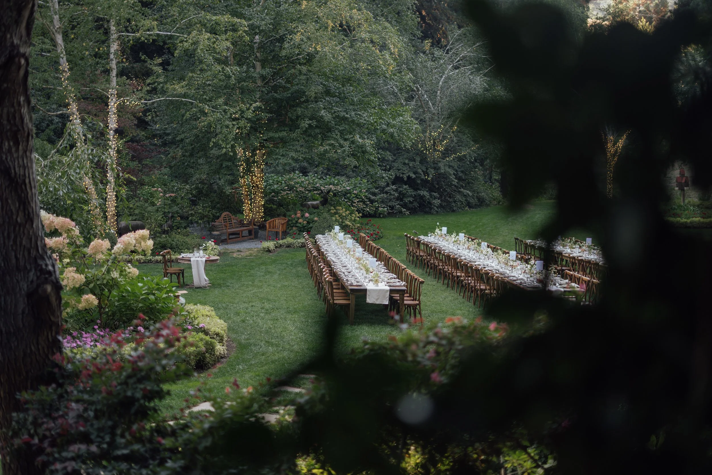 Outdoor garden setting with long banquet table and chairs, decorated with white tableware, amidst lush greenery and trees with string lights, suggesting a setup for an event or celebration.