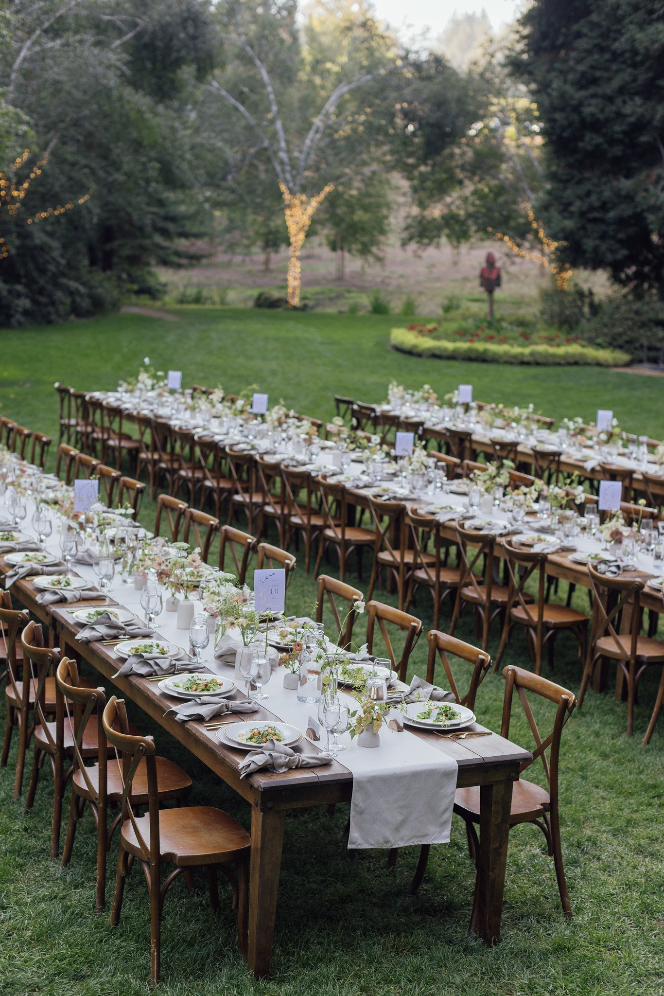 Long banquet tables set with white tablecloths, plates, glasses, napkins, and floral centerpieces in a garden with trees and string lights.