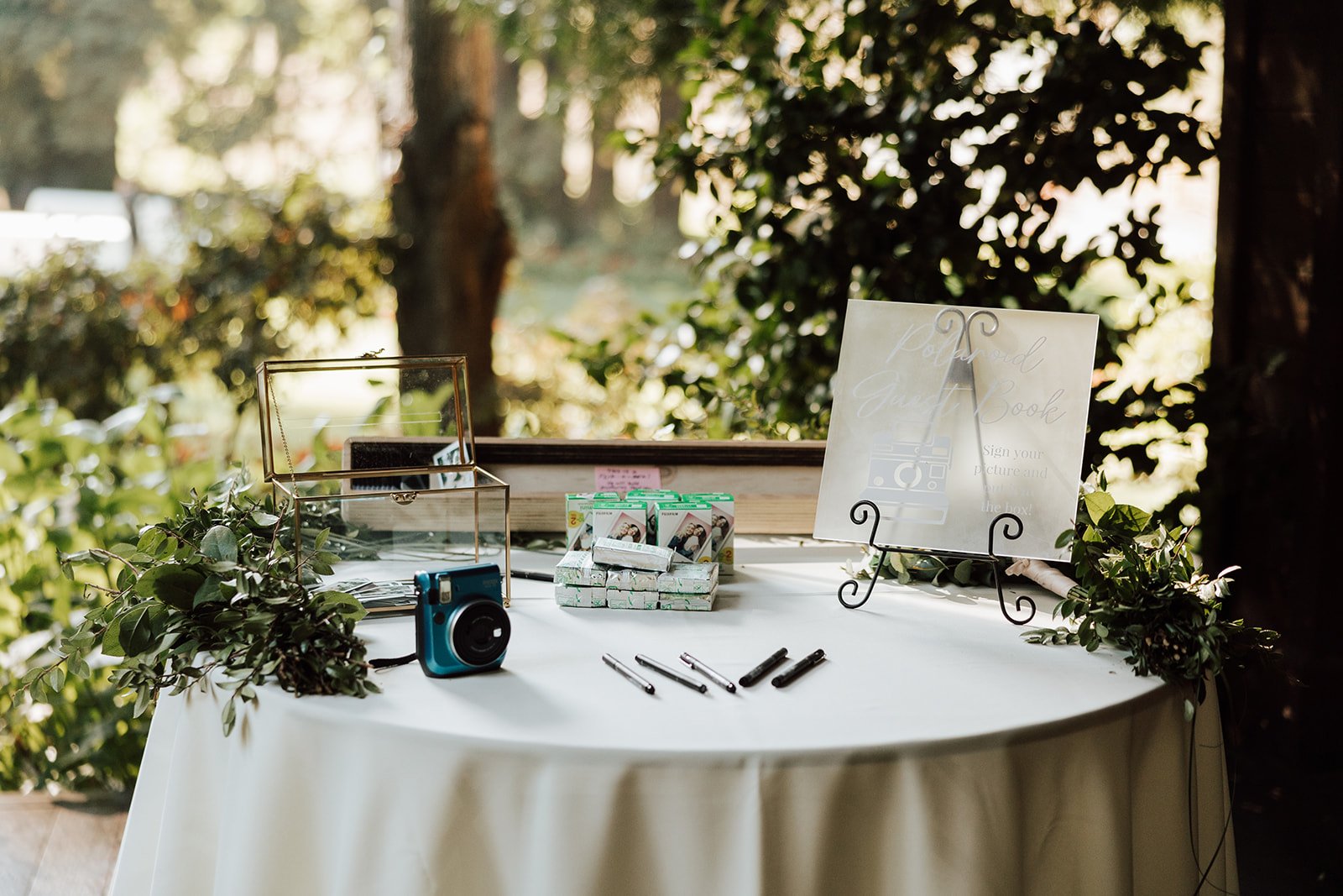 Decorative table for a guestbook and photo sign-in at an outdoor event, with a box of film or photo paper, pens, and greenery, set in a natural wooded environment.