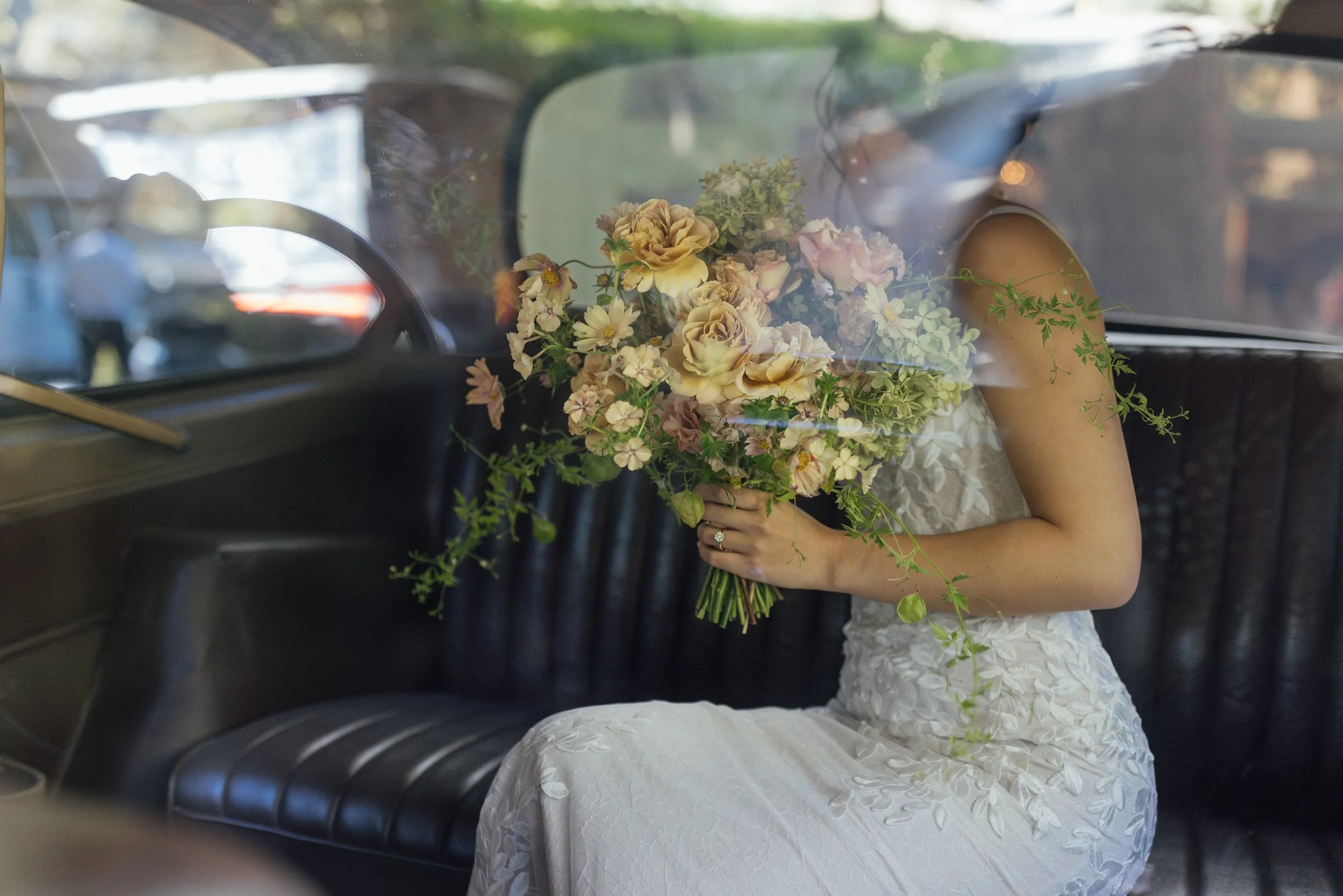 A woman in a white lace dress sitting inside a vintage car, holding a bouquet of pink, peach, and cream flowers with greenery, viewed through the car's window.