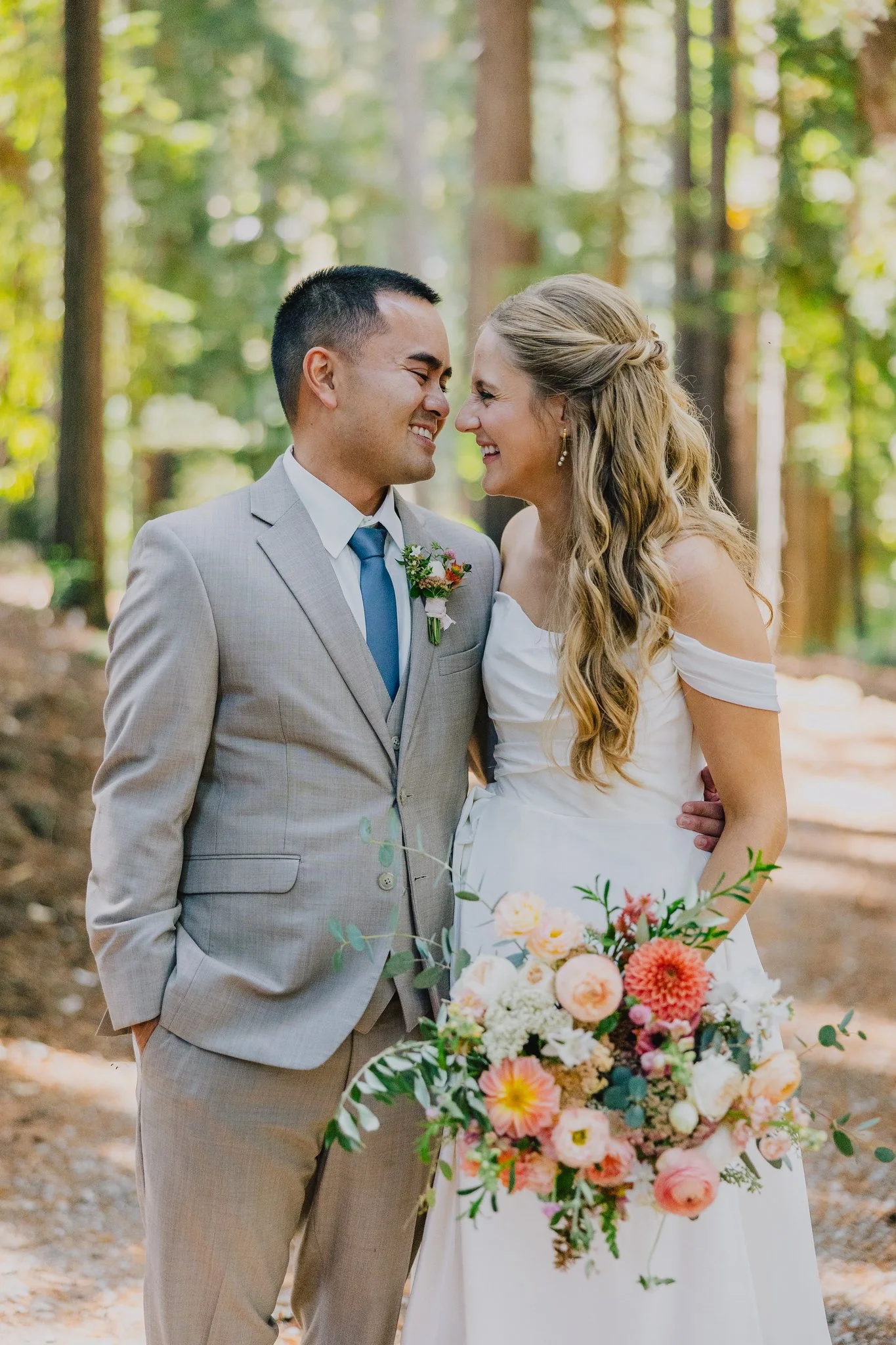 A newlywed couple smiling and close together in a forest, with the bride holding a large bouquet of flowers.