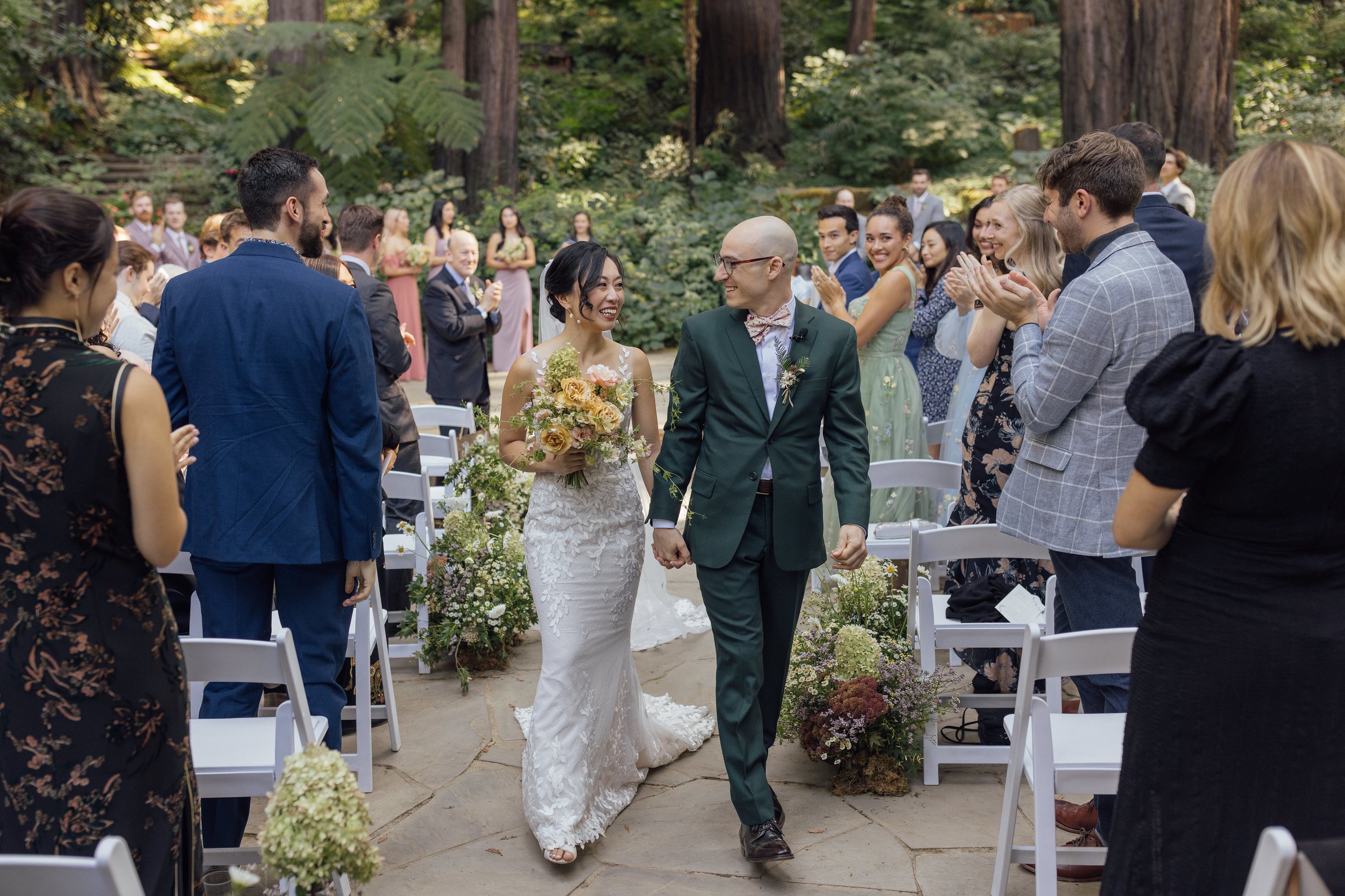 Bride and groom holding hands walking down the aisle at an outdoor wedding, surrounded by smiling guests in colorful attire.