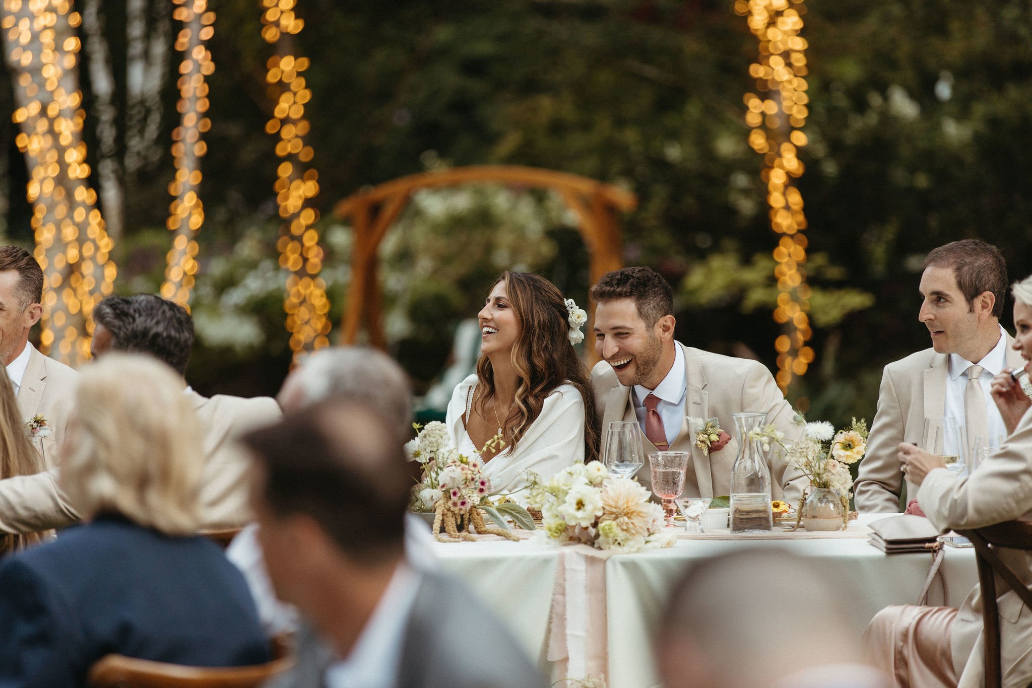 Bride and groom sitting at a decorated outdoor wedding reception table, smiling and laughing, surrounded by guests, with string lights in the background.