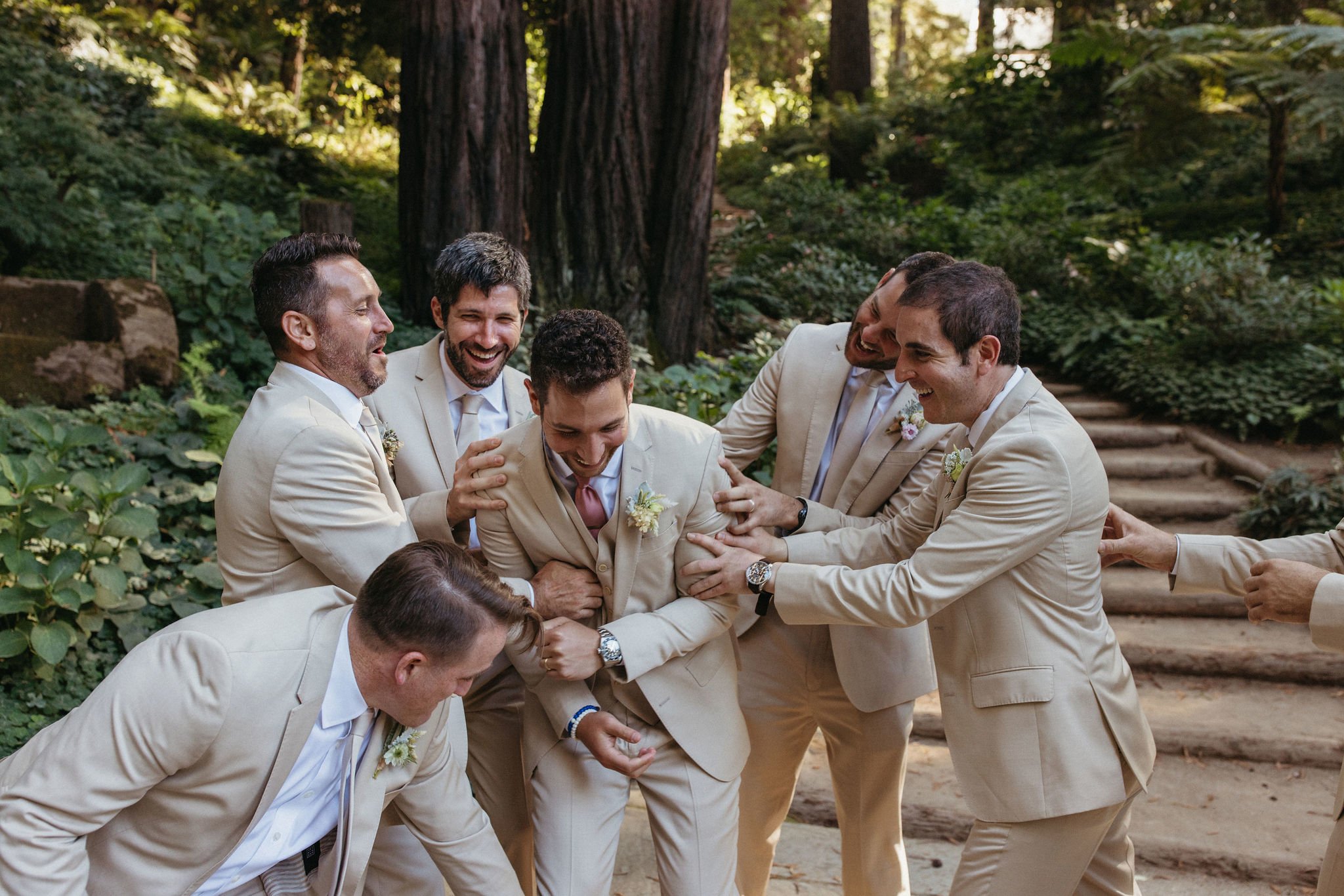 A group of men dressed in beige suits laughing and helping each other in a forested outdoor setting, celebrating a joyful moment.