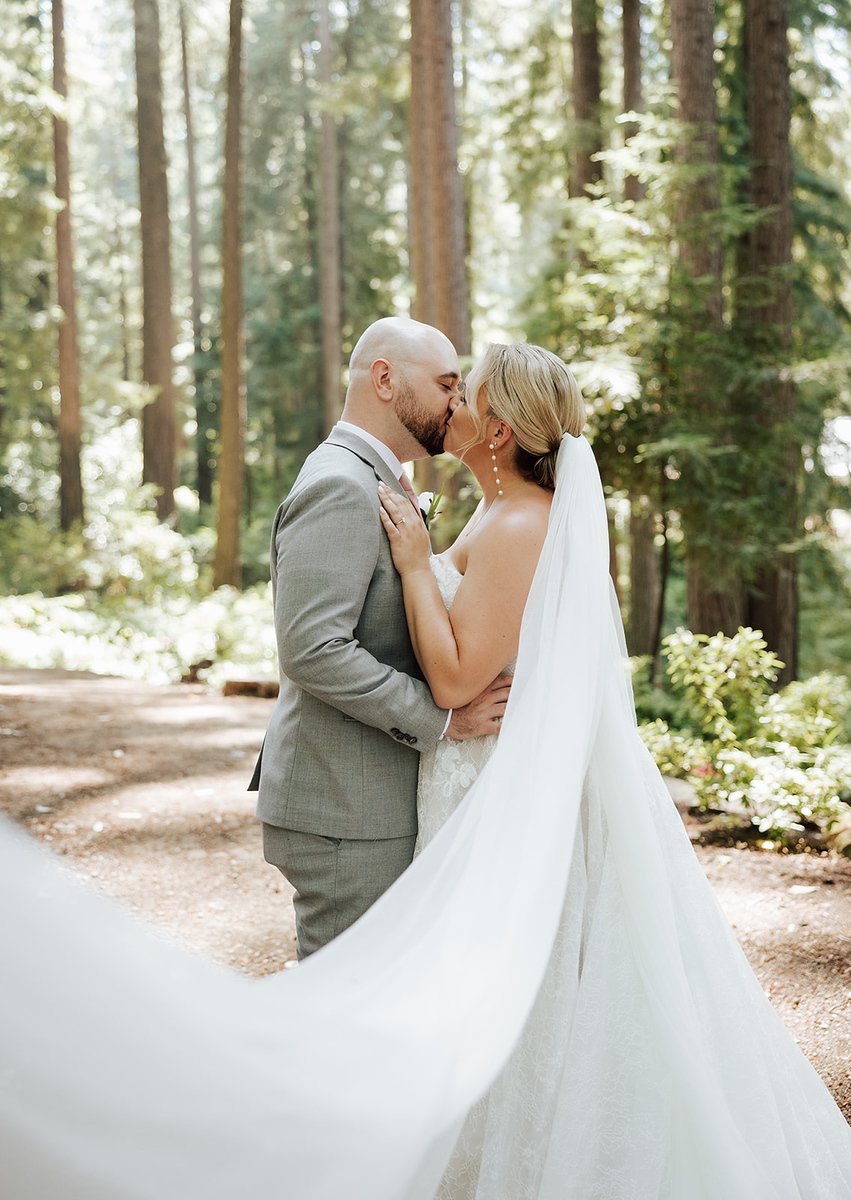 A bride and groom sharing a kiss in a forest setting during their wedding, with trees and sunlight in the background.
