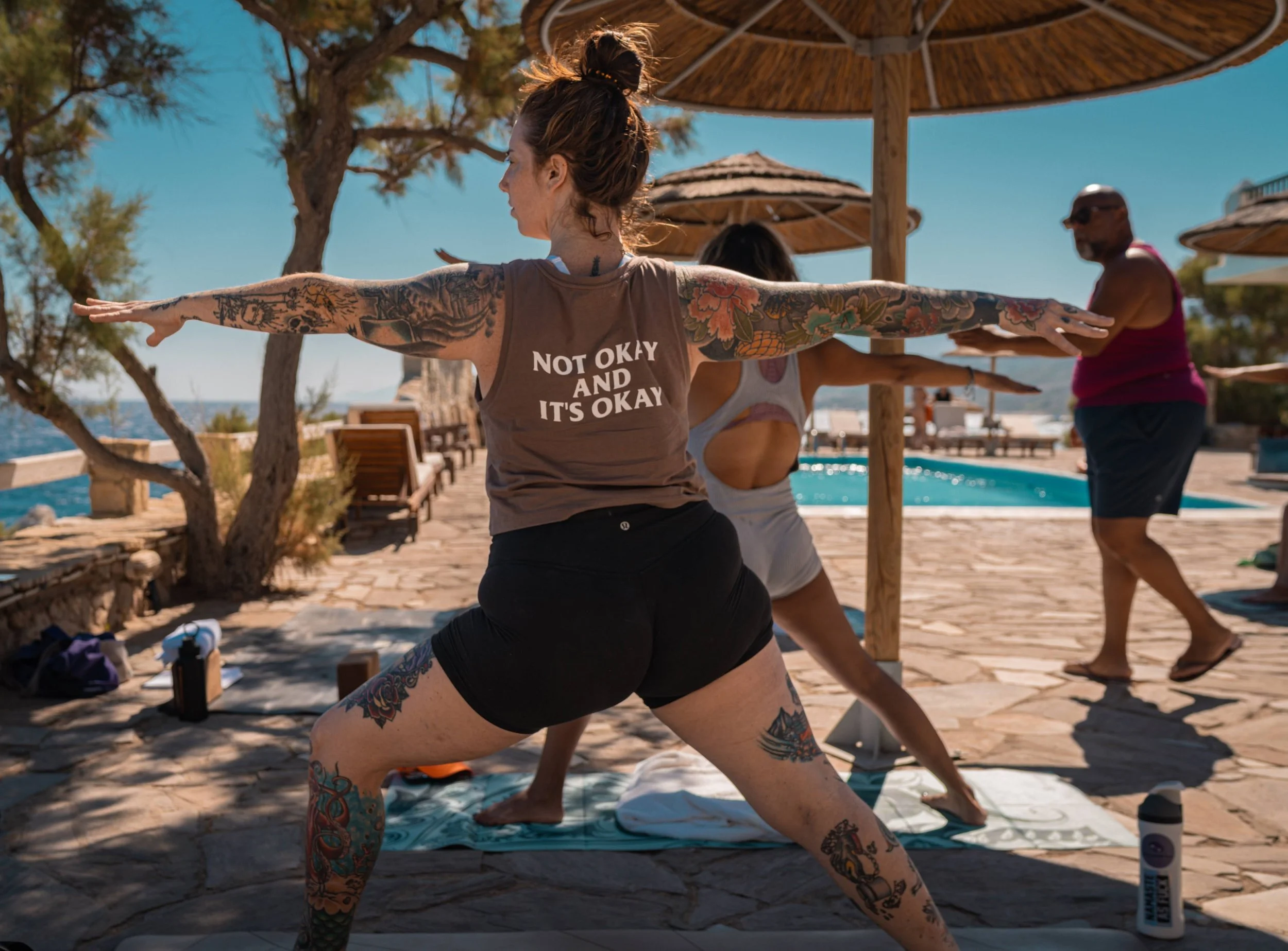 A woman with tattoos practicing yoga outdoors near a pool under a large umbrella, with other people, trees, and bright blue sky in the background.