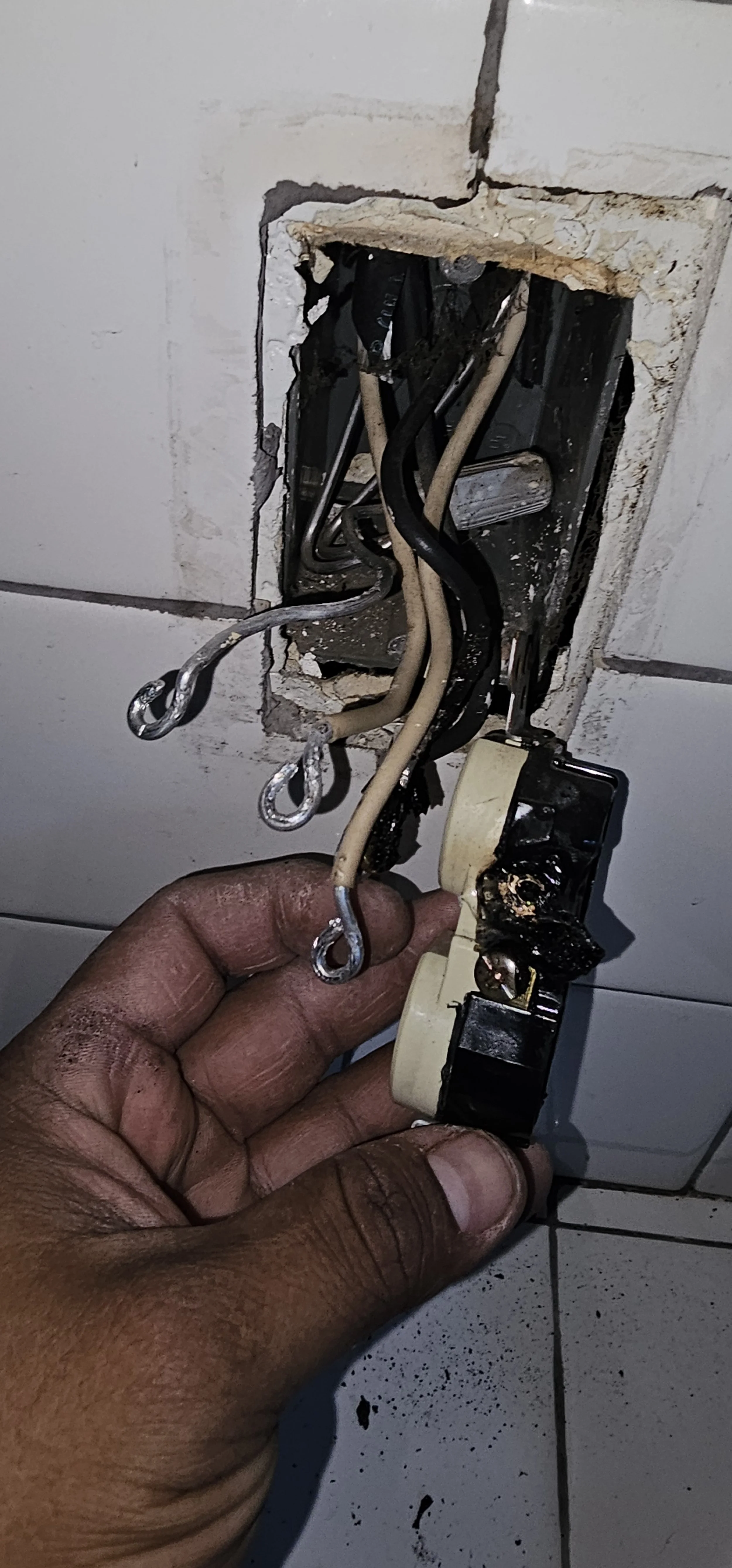 A person's hand holding an old, burned-out electrical wiring and switch in a wall opening, with wires exposed and damaged.