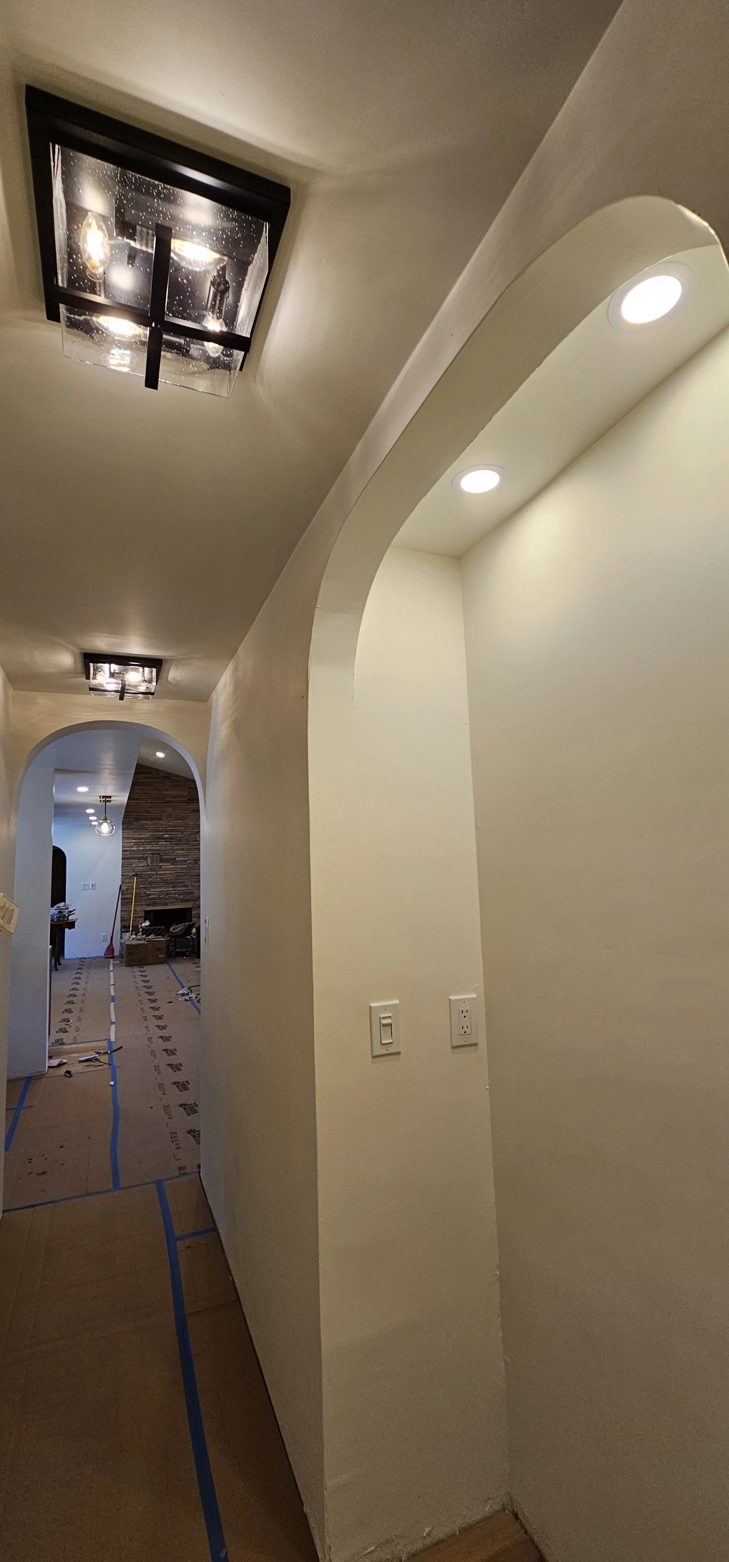 Interior hallway of a house with recessed lighting, a decorative arched wall opening, switches on the wall, and a view of a room with a brick fireplace in the background.