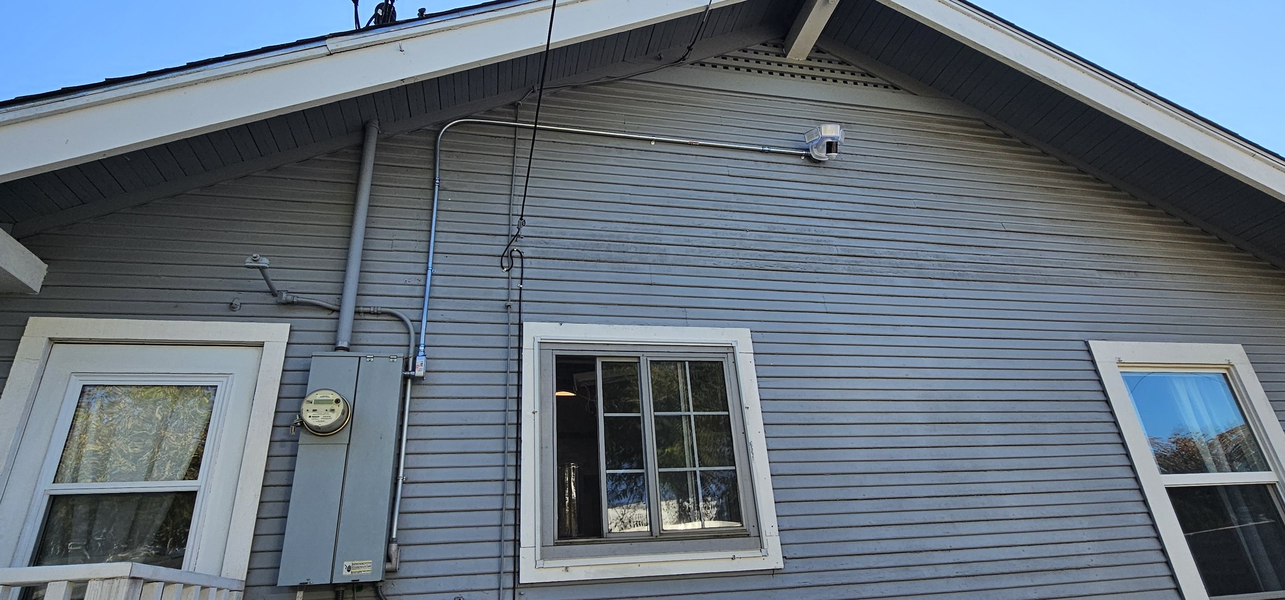 Close-up of the gray house's exterior with two windows, an electric meter, and electrical conduit on the siding, under a blue sky.