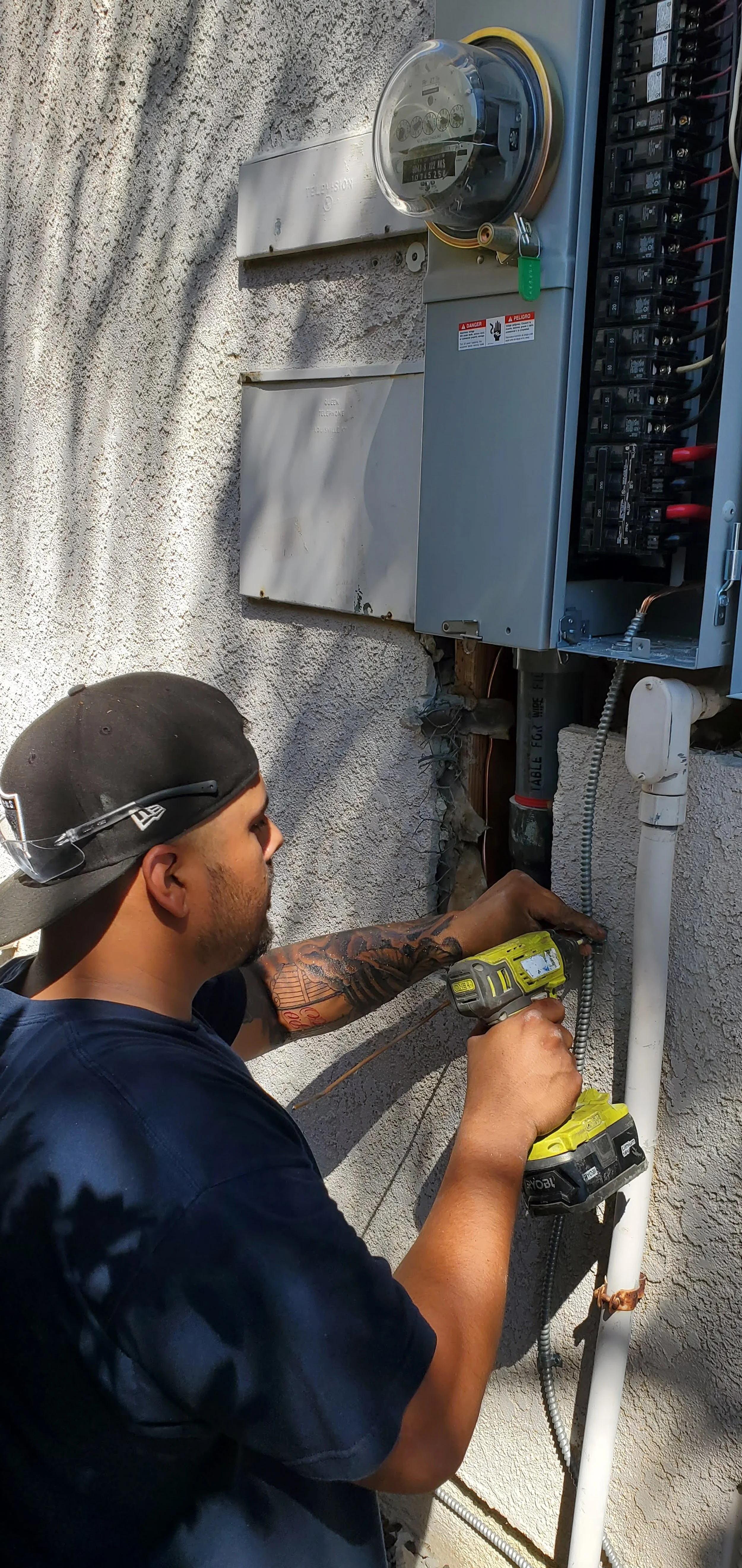 A man working on an electrical panel outside a building, using a cordless drill.